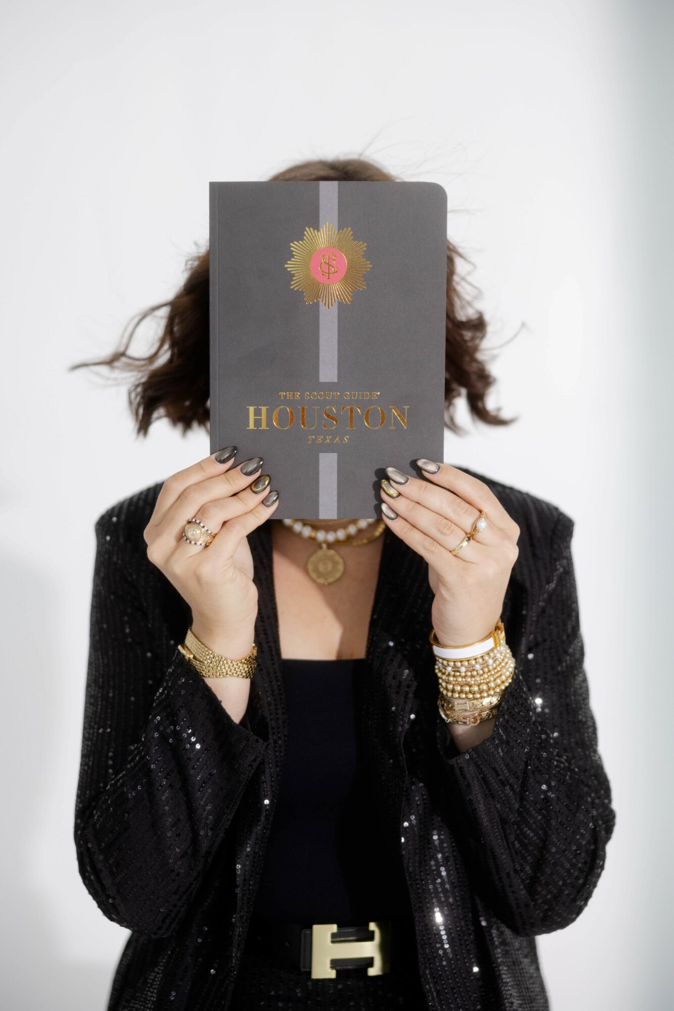 Person holding a book in front of their face, wearing a black outfit with jewelry, against a plain background.