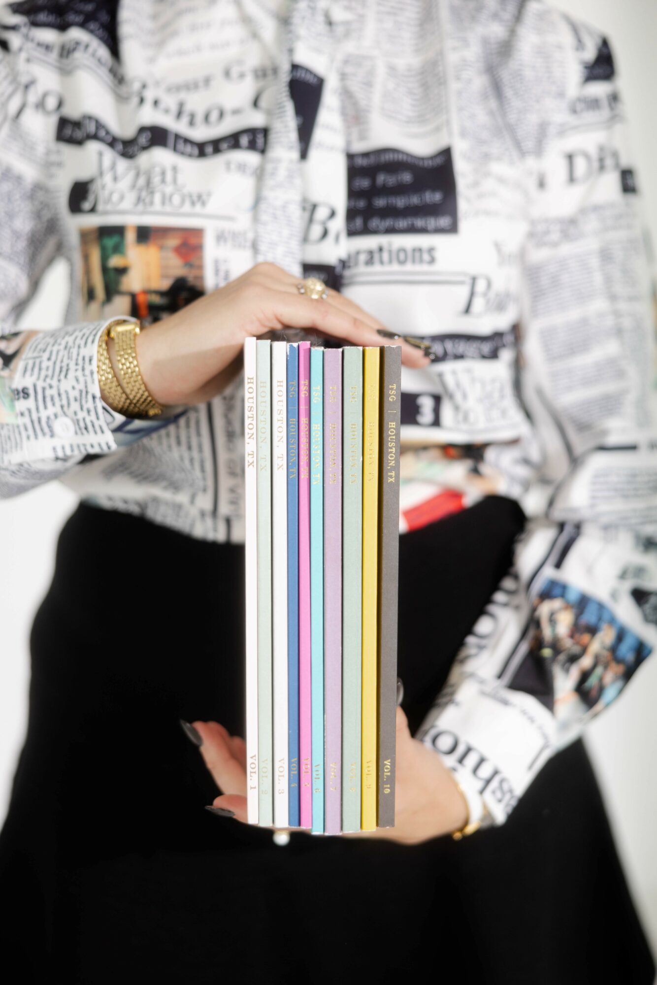 Person holding a colorful stack of books, wearing a newspaper-patterned jacket and gold bracelet, with a blurred background.