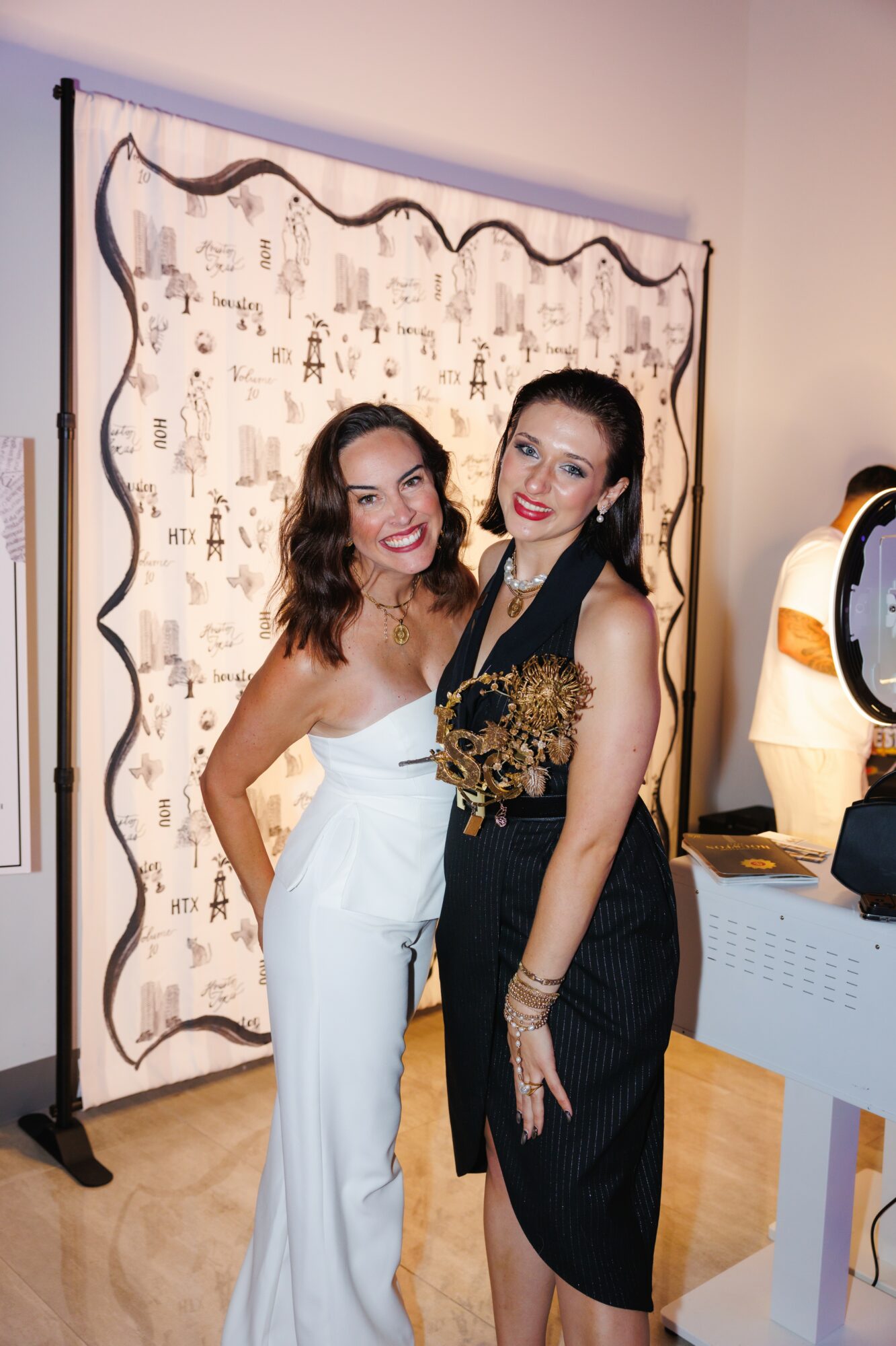 Two women in elegant outfits pose together indoors, smiling, with a decorative backdrop behind them.