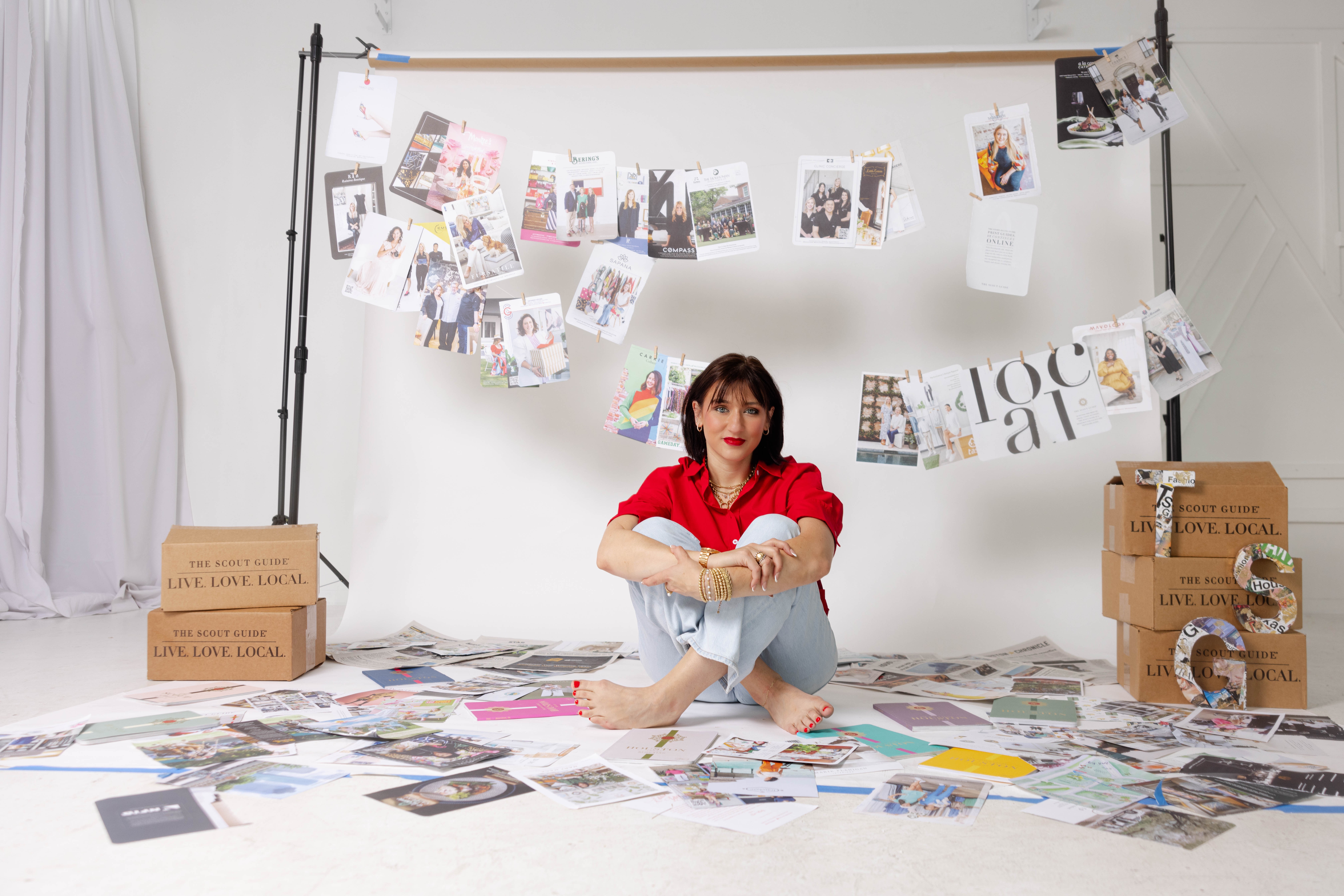 Woman sitting on floor with photos and papers, decorated backdrop, cardboard boxes, and hanging photos behind her.