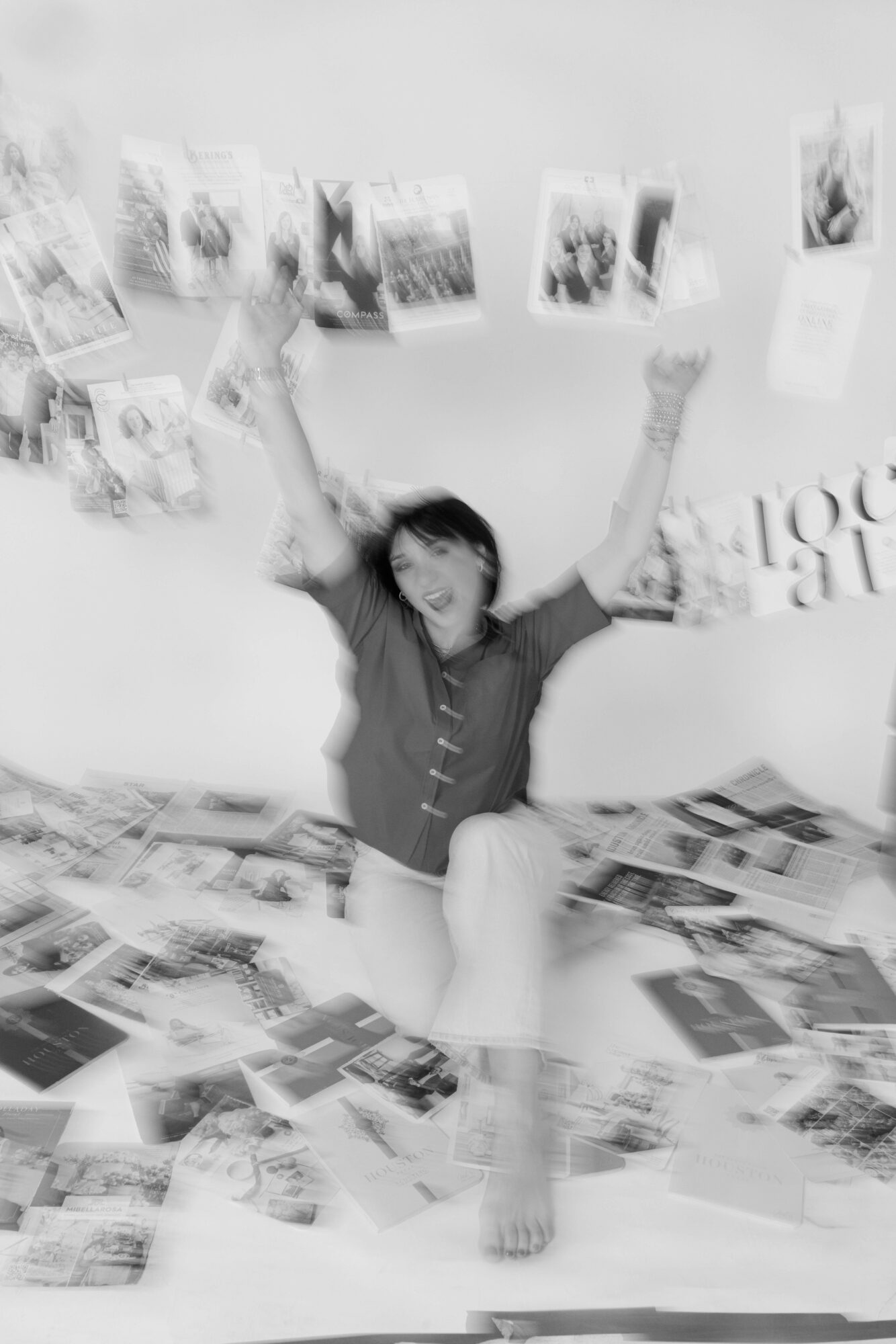 Person sitting with arms raised, surrounded by photos and magazines on wall and floor, smiling.