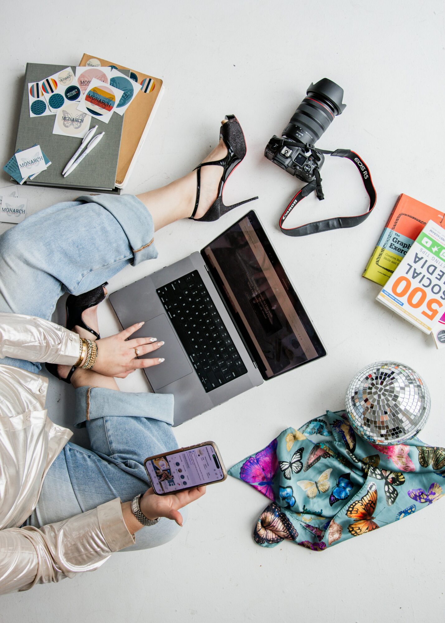 Person using laptop, holding smartphone, with camera, notebook, and colorful scarf on white surface.