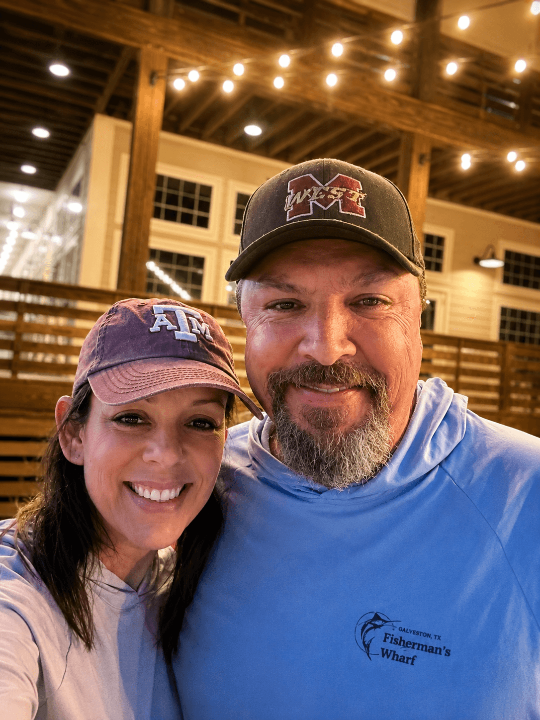 A smiling man and woman taking a selfie indoors with string lights and wooden beams overhead.