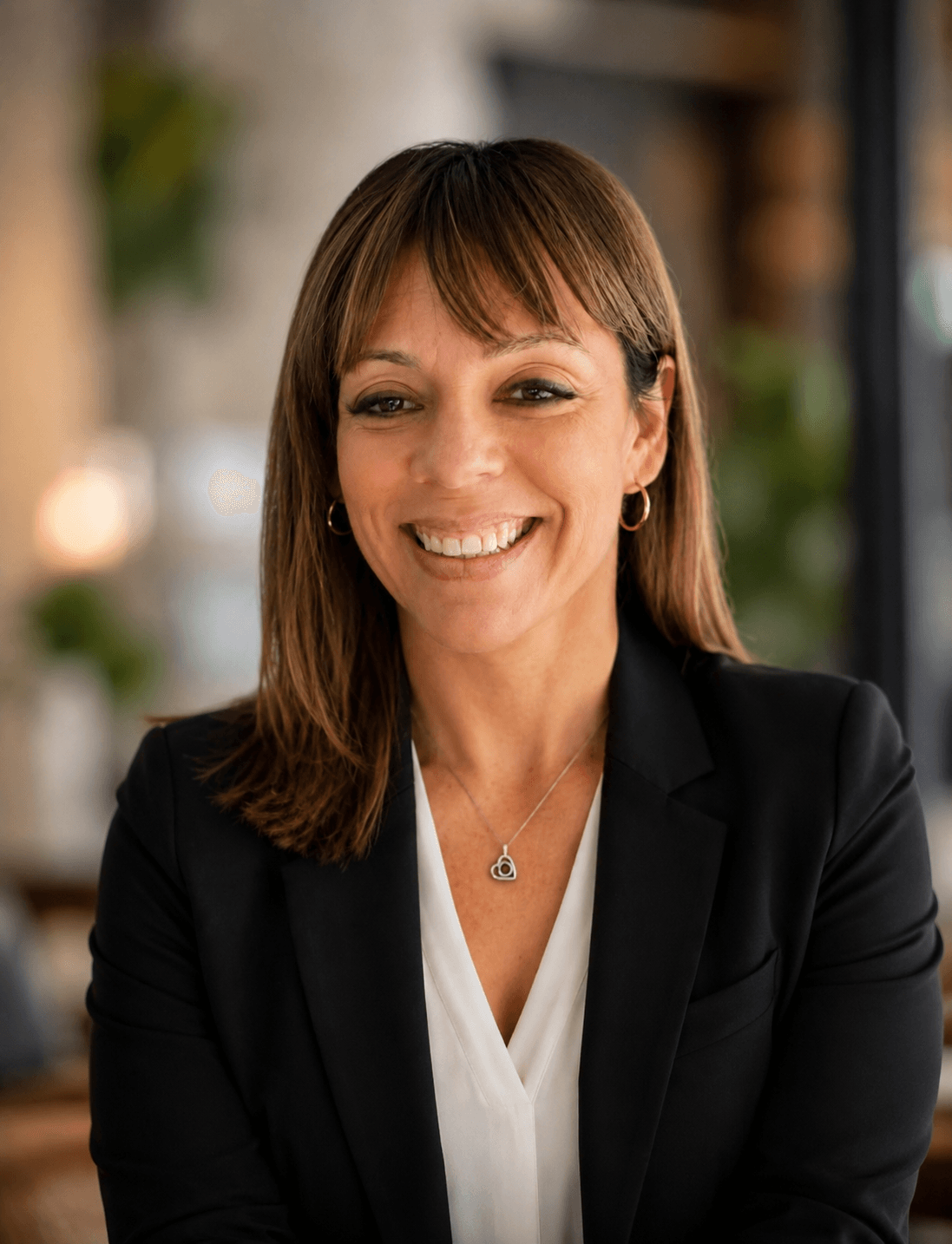 Smiling woman with shoulder-length brown hair, wearing a black blazer and white top, indoors with blurred background.