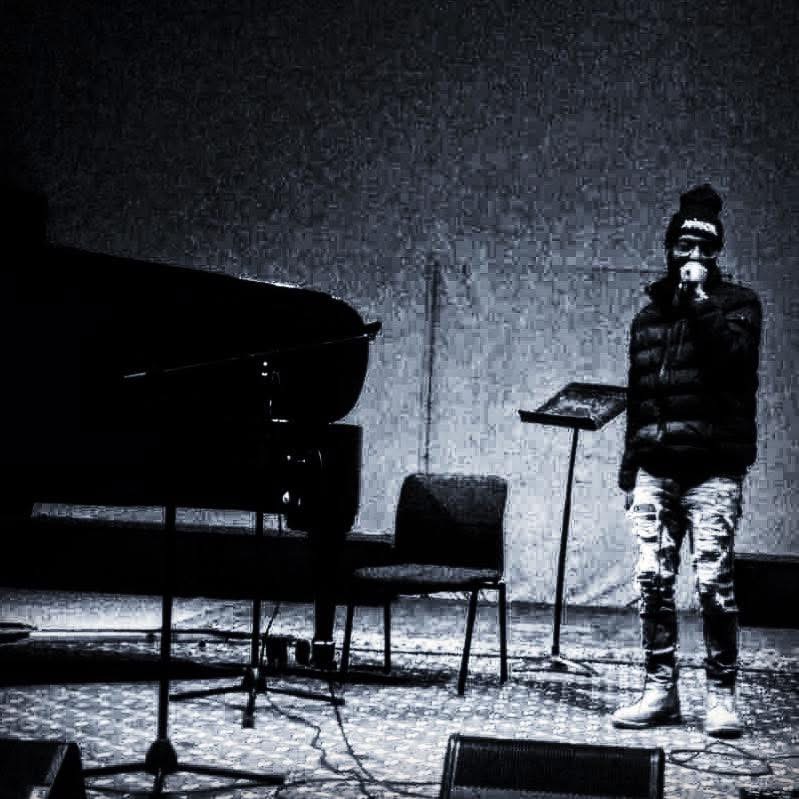 Person standing on stage with a microphone, a piano, and chairs, in a dark setting, possibly during a performance or rehearsal.