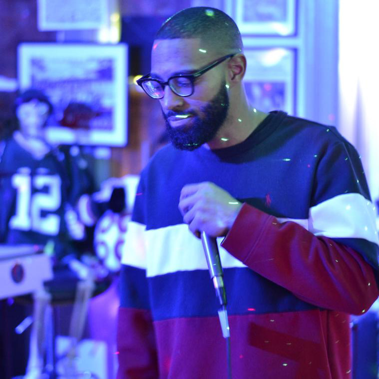 Man with glasses and beard holding a microphone, wearing a color-blocked sweatshirt, in a room with framed pictures. Word count: 24