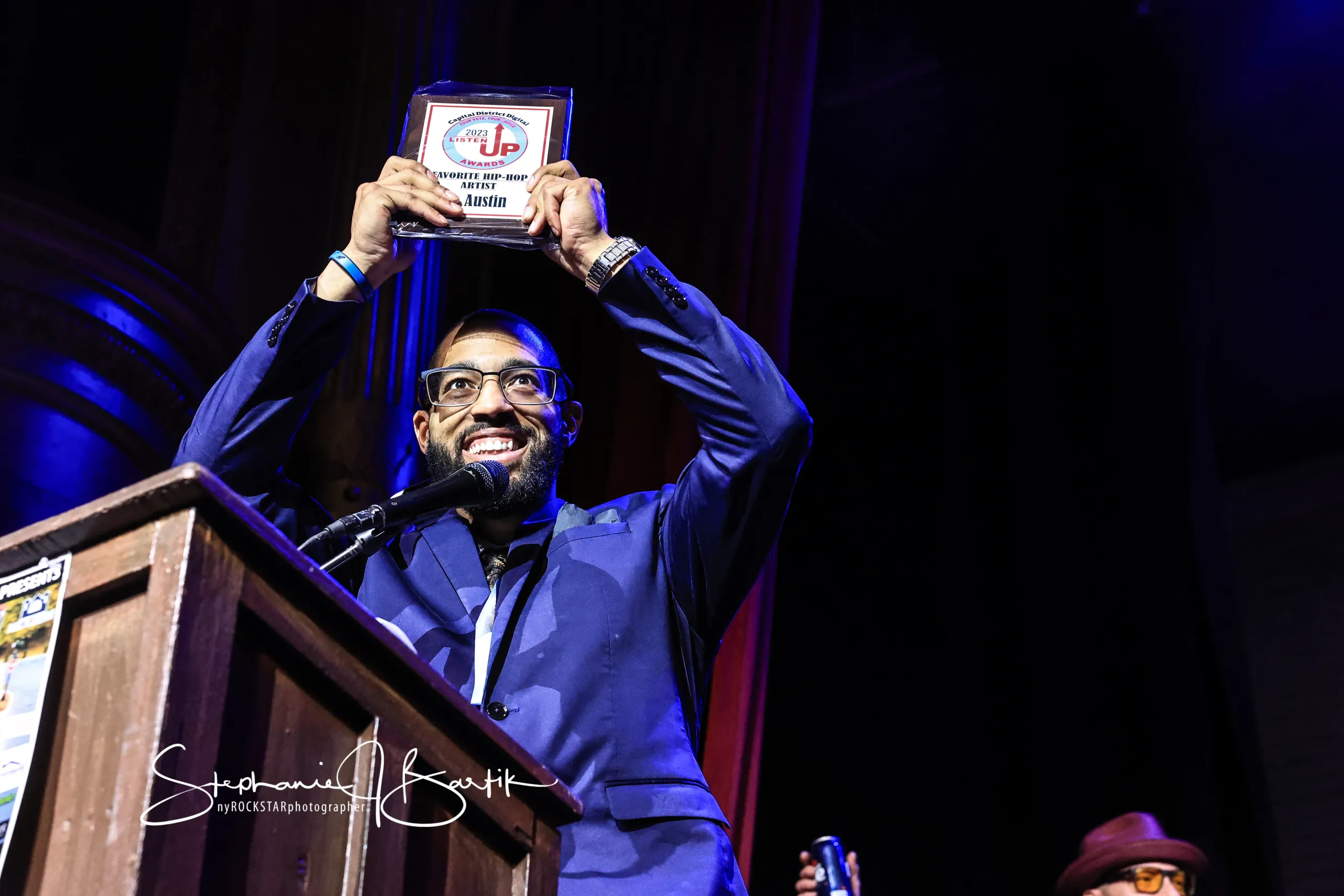 Man with glasses and beard holding an award above his head on stage, smiling, with a podium in front, dark background.