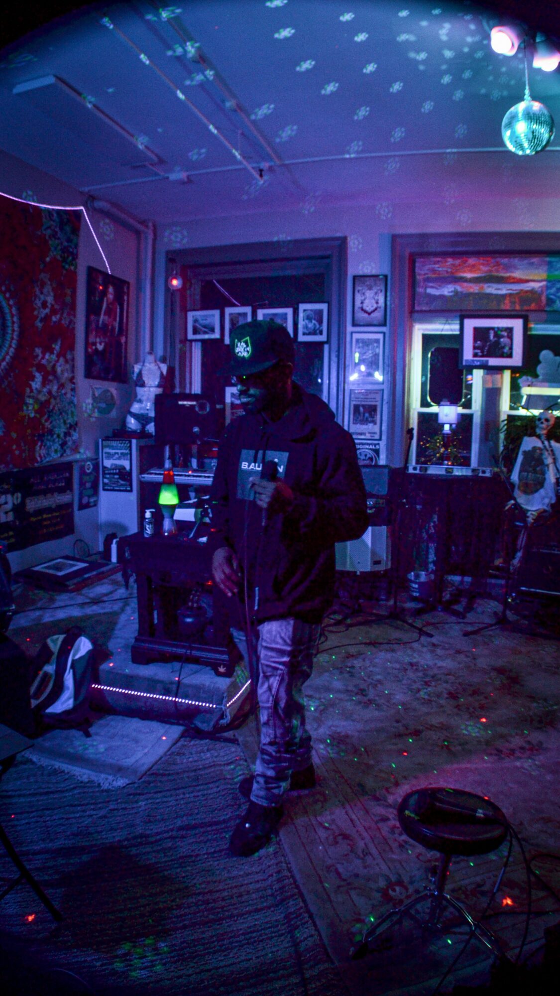 Person standing in a dimly lit room with colorful artwork and electronic equipment, illuminated by blue and purple lights.