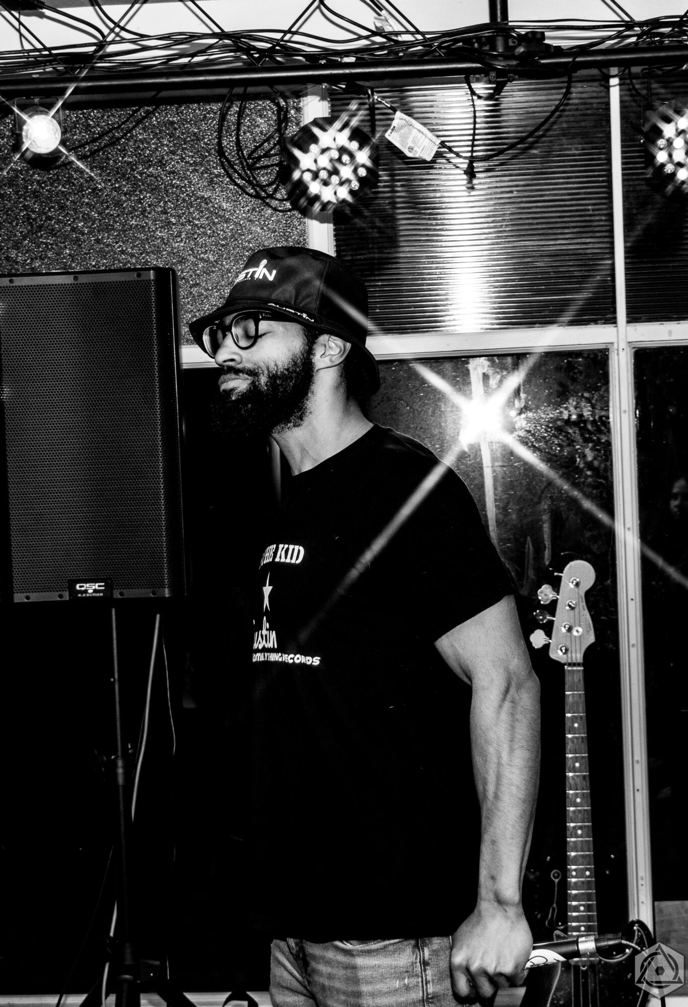 Man with glasses and a hat stands near a speaker, with a guitar and bright light in the background.