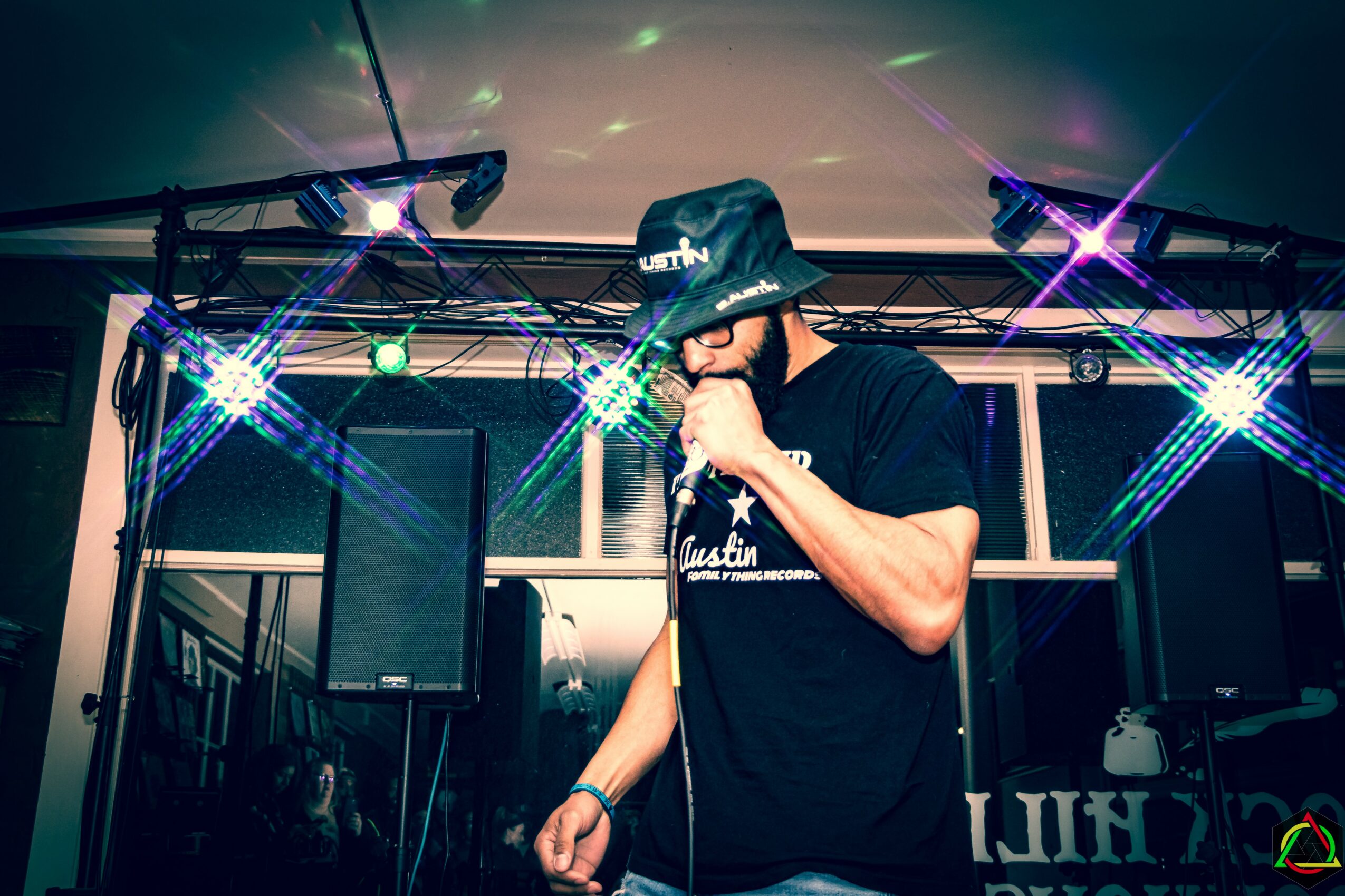 Person wearing a hat and black T-shirt performing with microphone on stage under colorful lights.