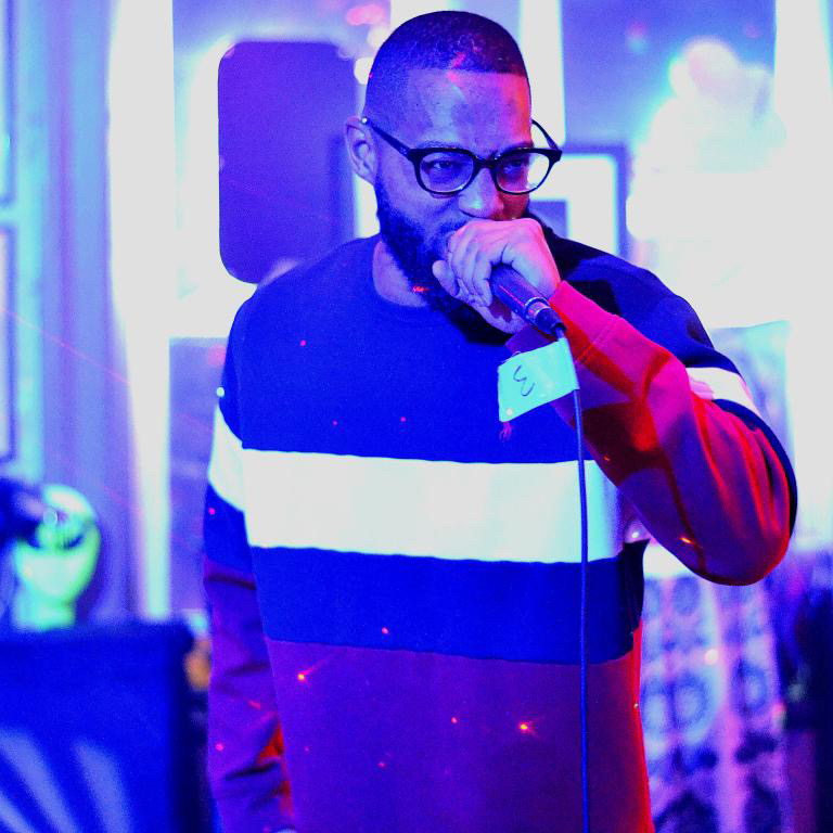 Man with glasses singing into a microphone in a colorful, illuminated setting.