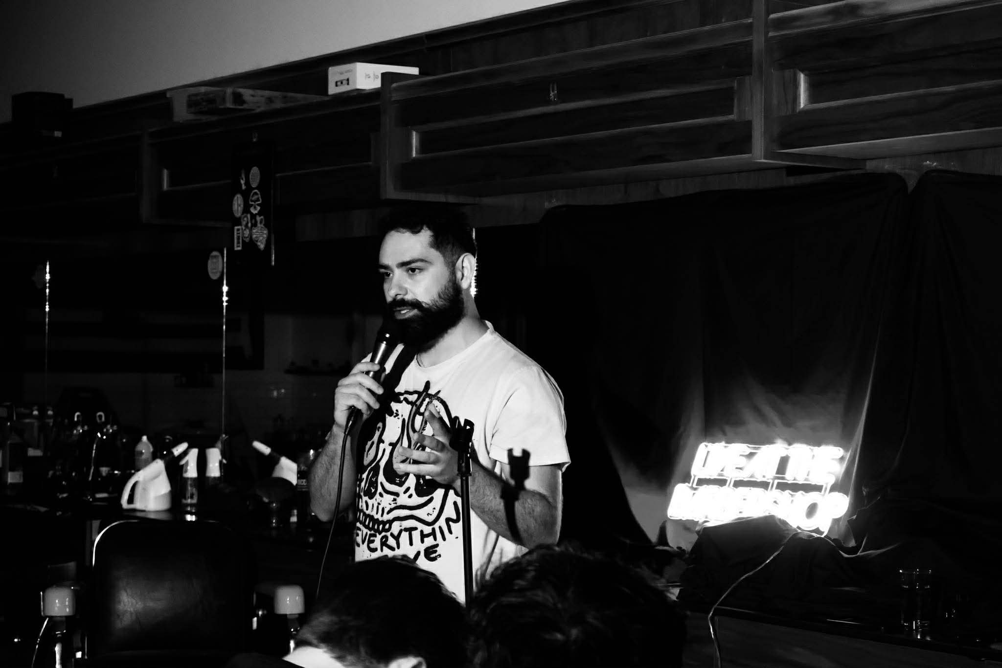 Man with a beard and tattooed arm speaking into a microphone on stage, with a dark background and audience.