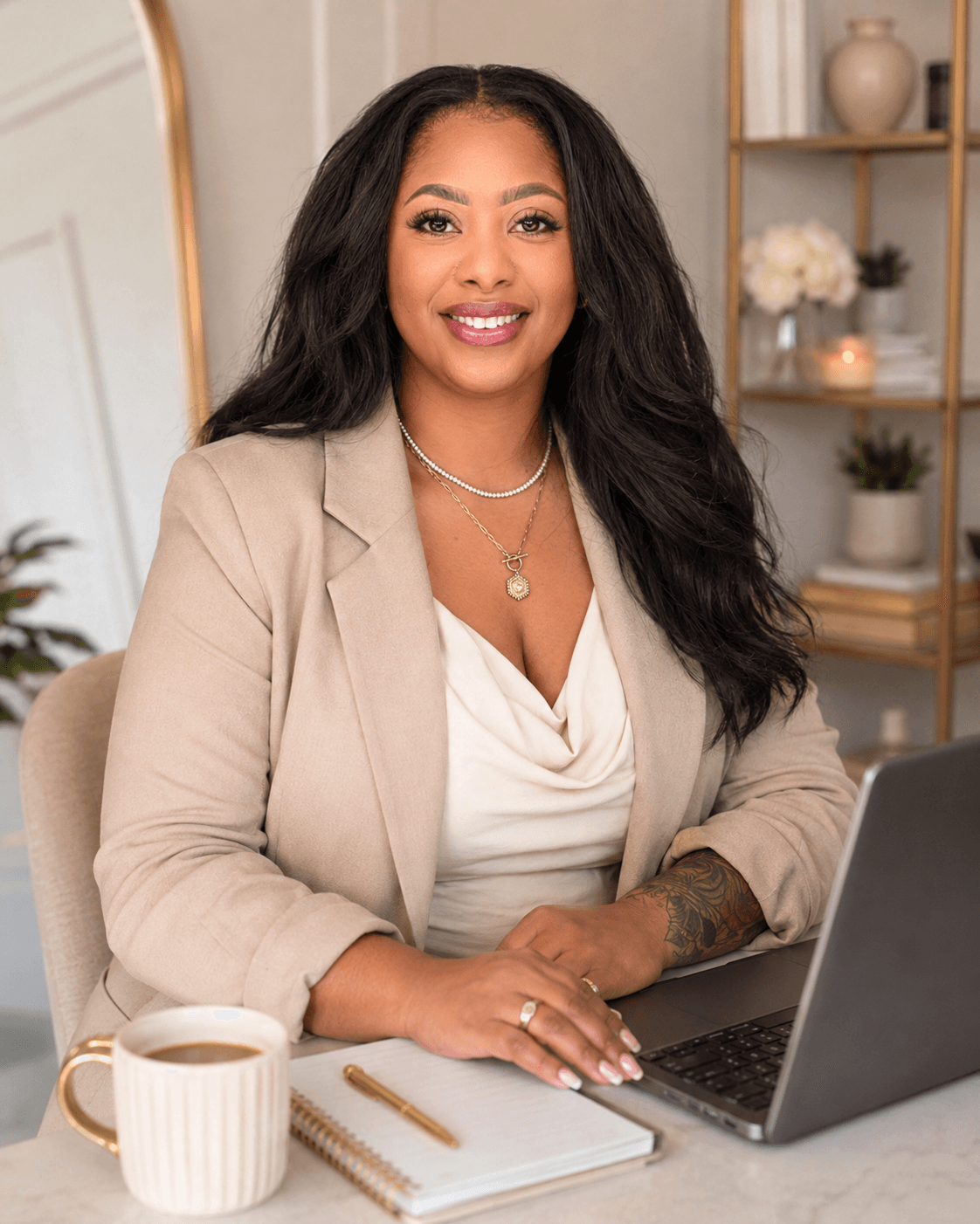 Woman with long dark hair smiling at camera, sitting at desk with laptop, notebook, and coffee mug, in a modern office.