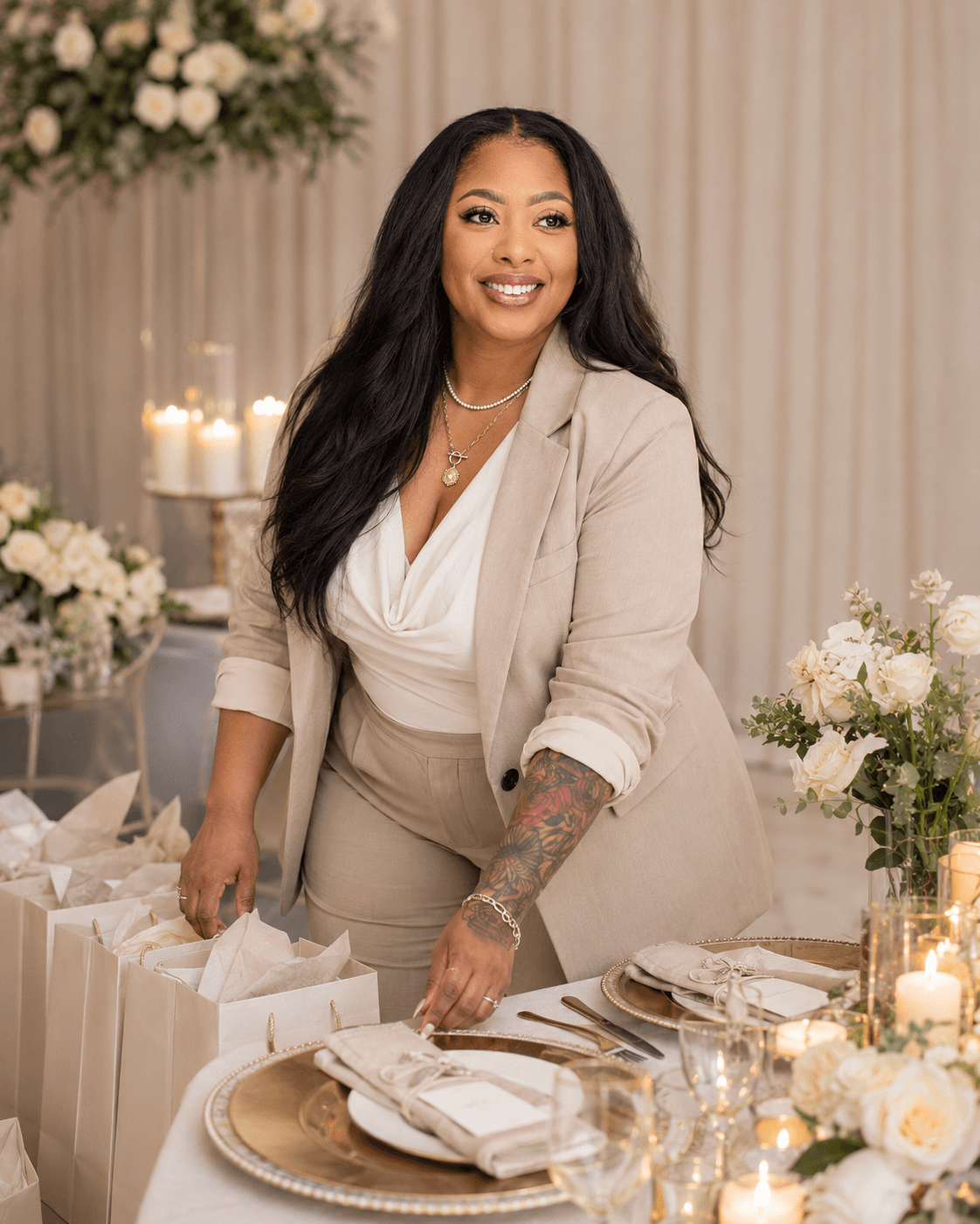 Woman in beige suit smiling at a decorated table with flowers and candles, holding a gift bag.