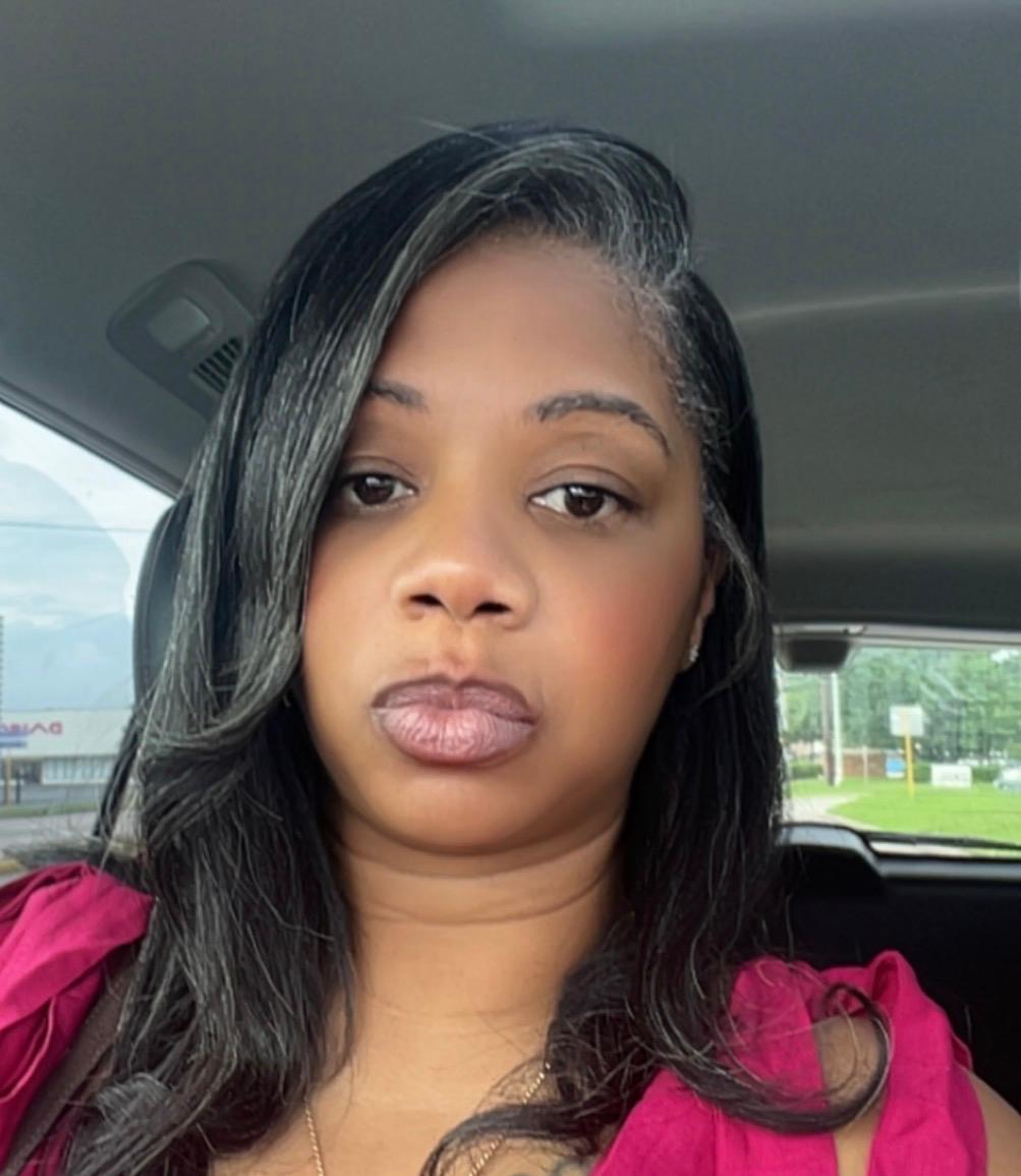 Woman with long black hair and pink top sitting in car, looking at camera, neutral expression.