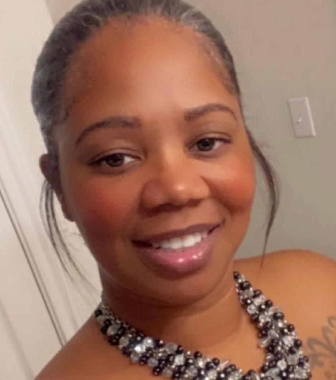 Close-up of a smiling woman with braided hair, wearing a beaded necklace, in a neutral background.