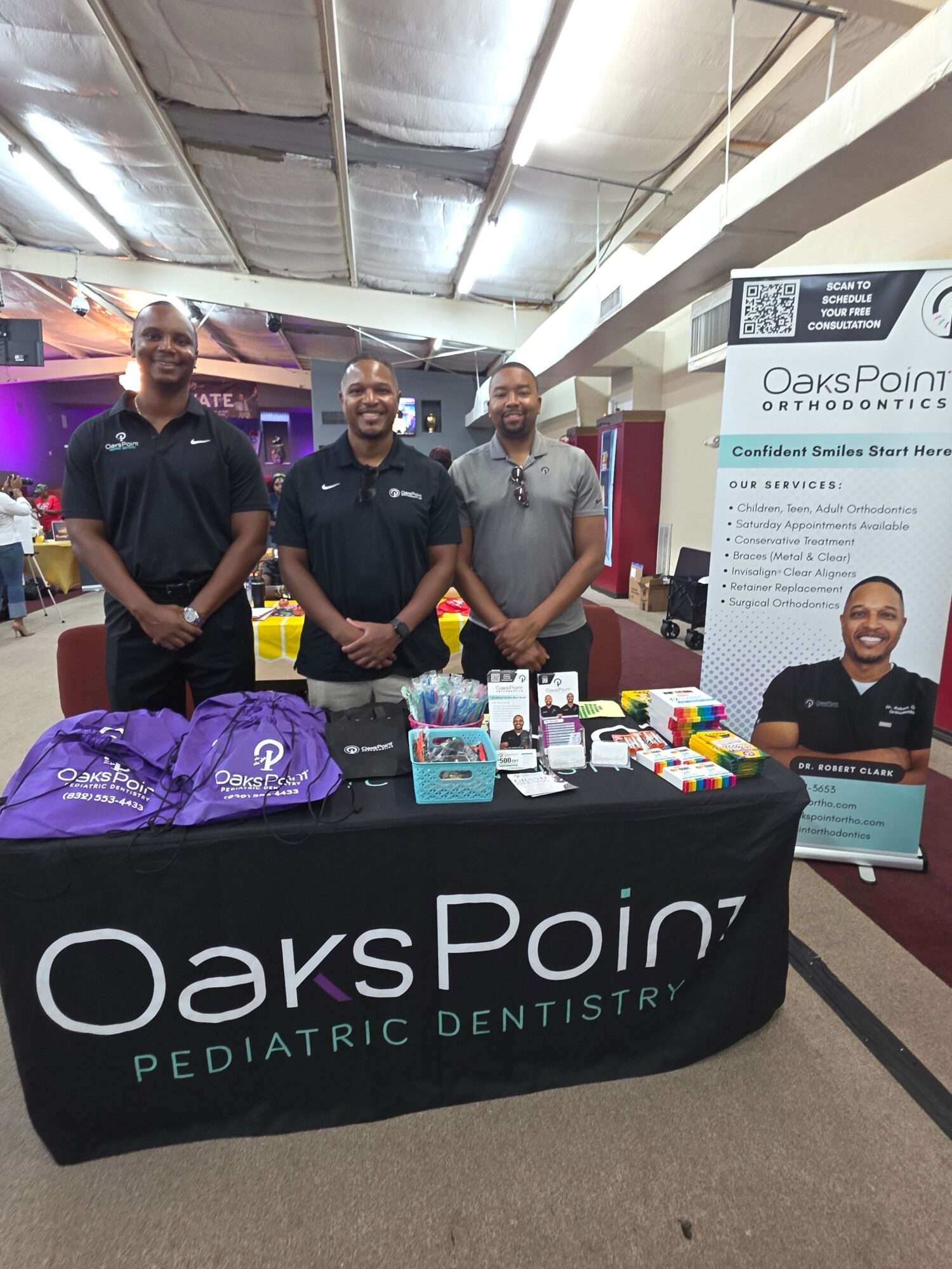 Three people standing behind a table with promotional materials at a dental event, with a banner in the background.