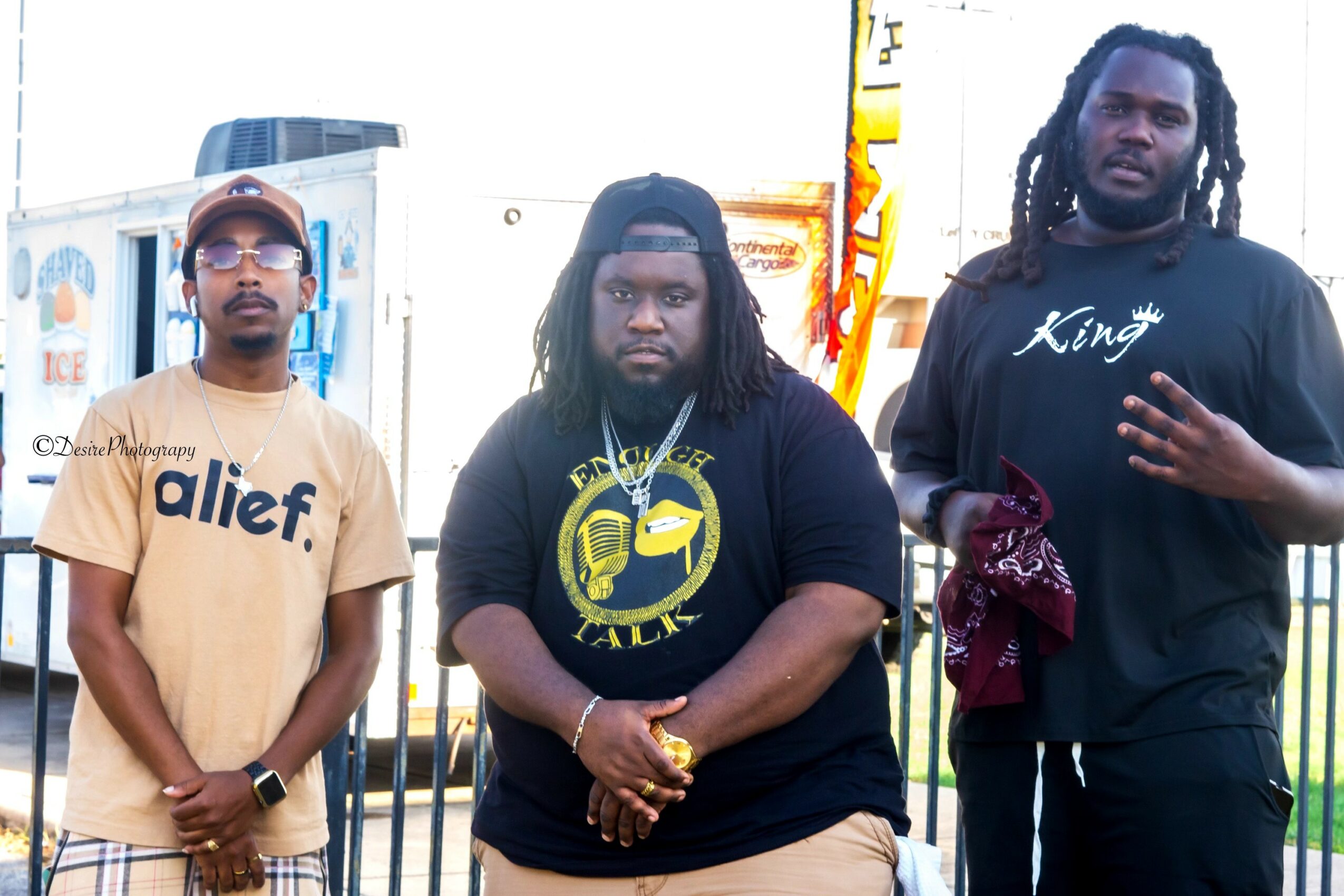 Three men standing outdoors with city buildings in the background, wearing casual clothing and accessories.
