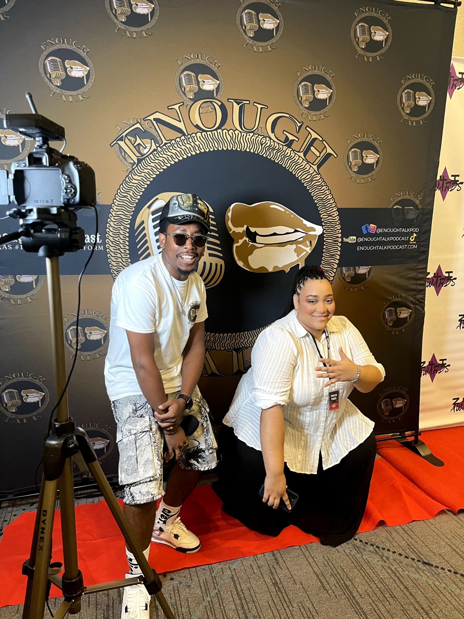 Two people posing in front of a black backdrop with gold text and logo, one standing and one kneeling, with a camera on a tripod nearby.