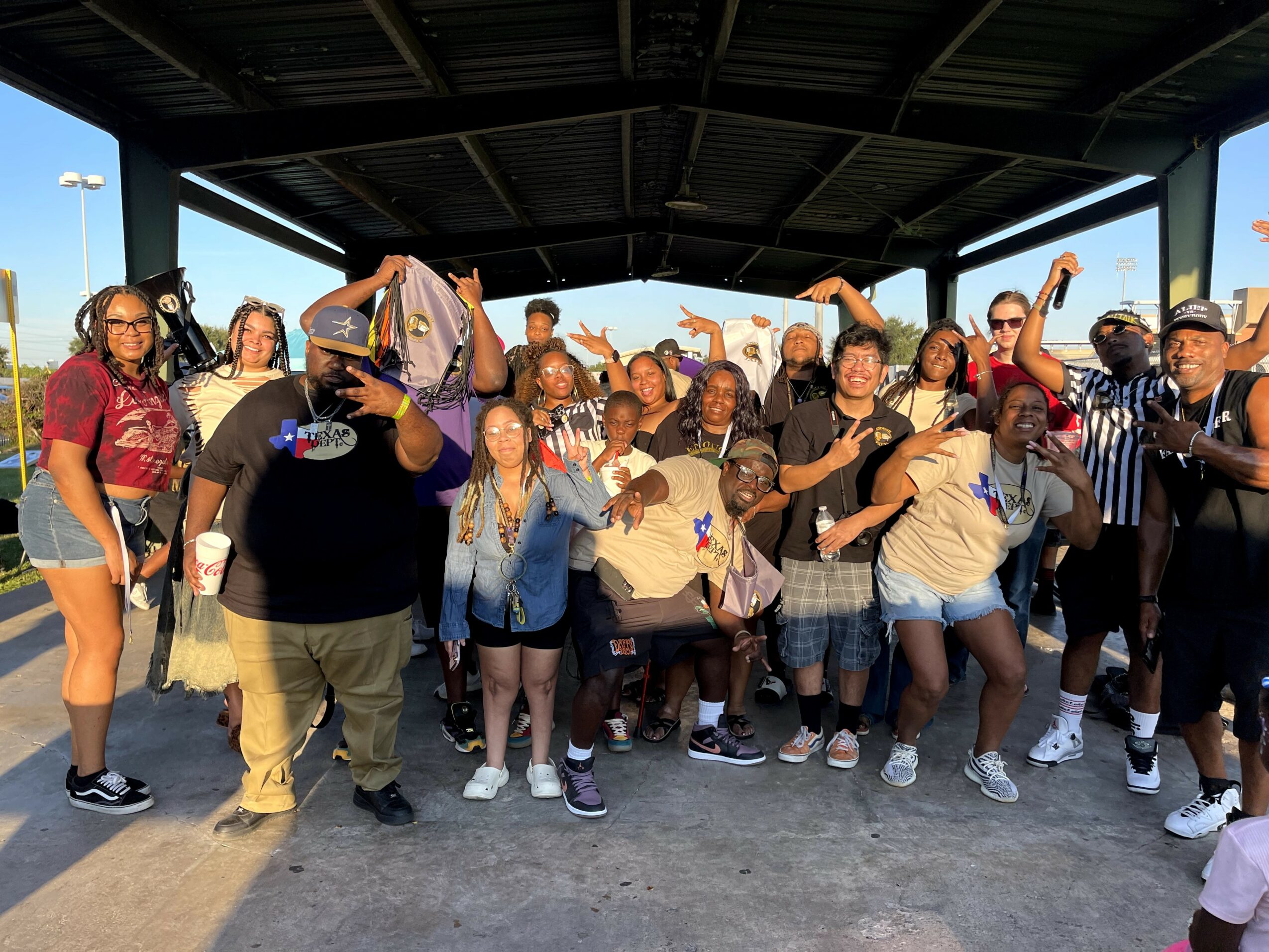 Group of diverse people posing outdoors under a shelter, some making peace signs and smiling.