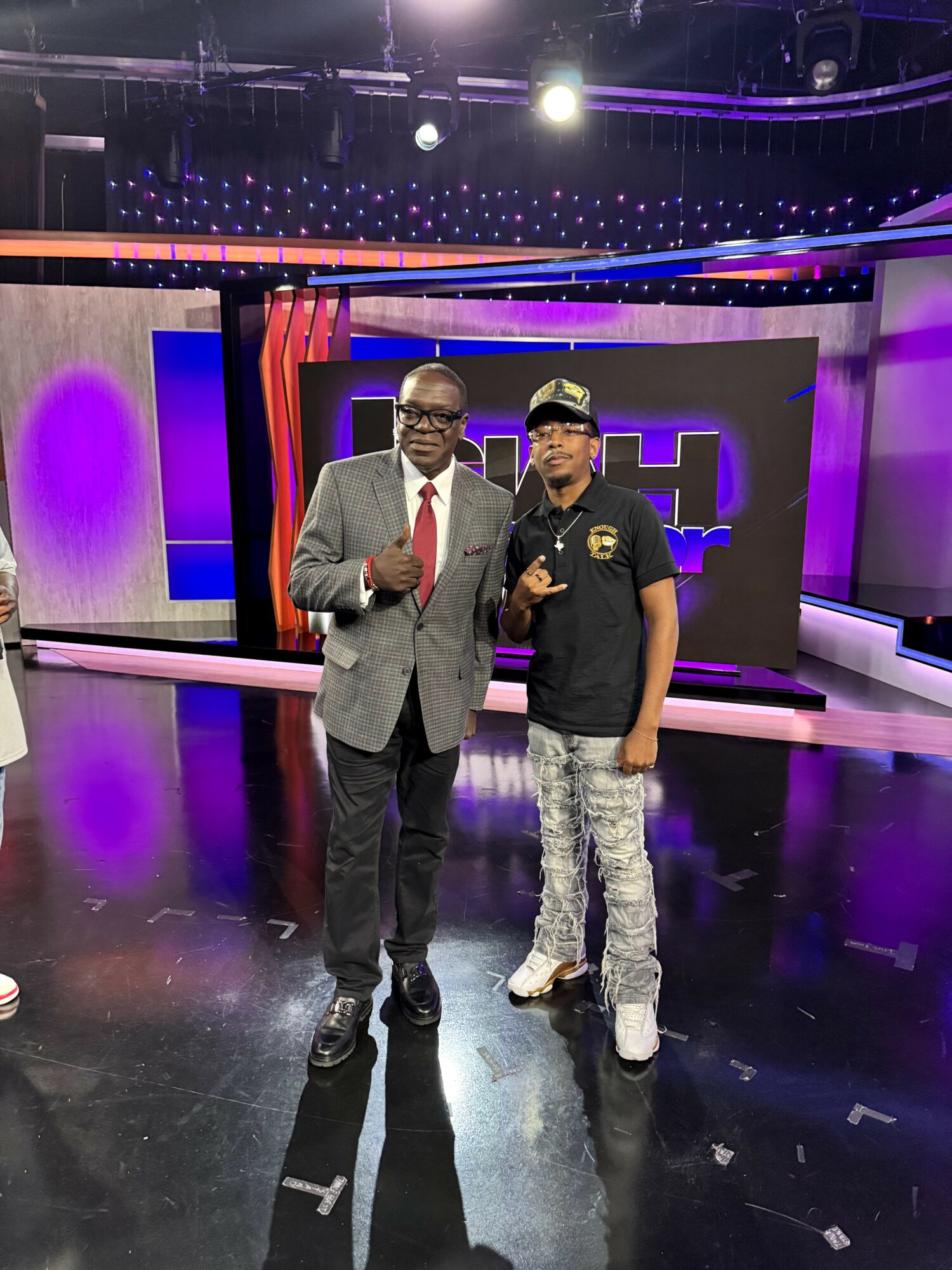 Two men standing on a television studio set with purple and black background, one in a suit, the other in casual clothing, both making peace signs.