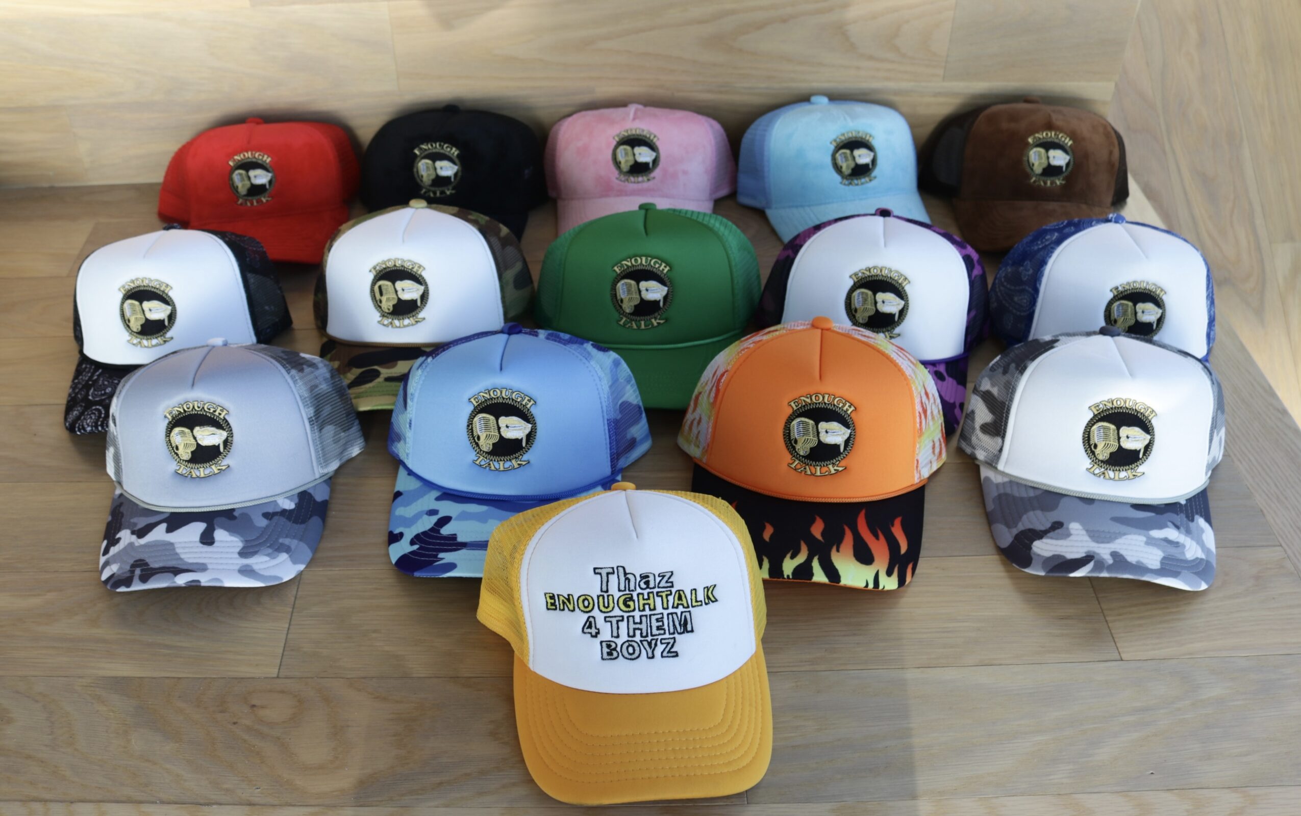 Collection of 15 colorful baseball caps arranged in three rows on a wooden surface, with a yellow and white cap in front.