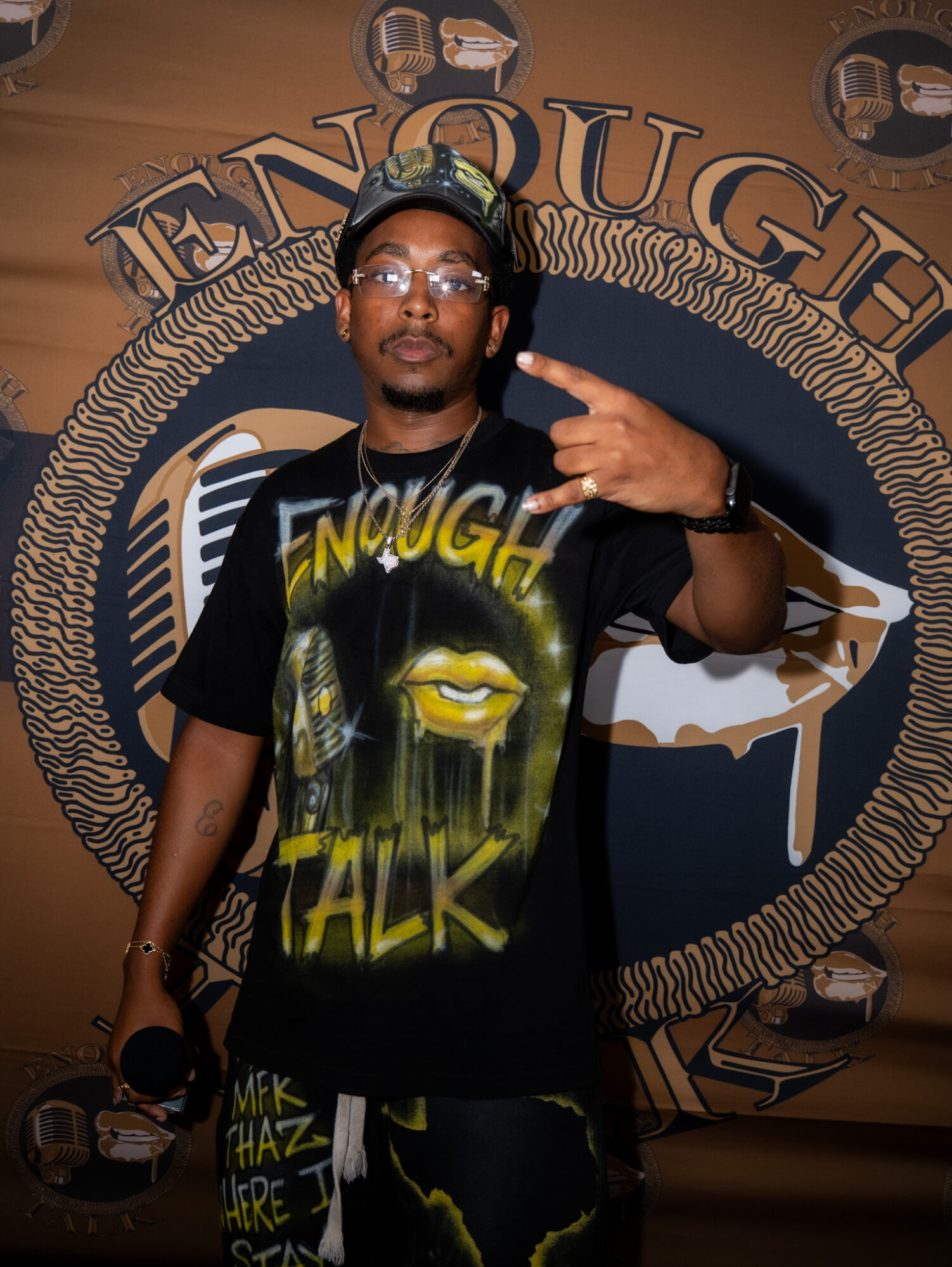 Young man wearing glasses and a cap, making a peace sign with his right hand, standing in front of a decorative background.