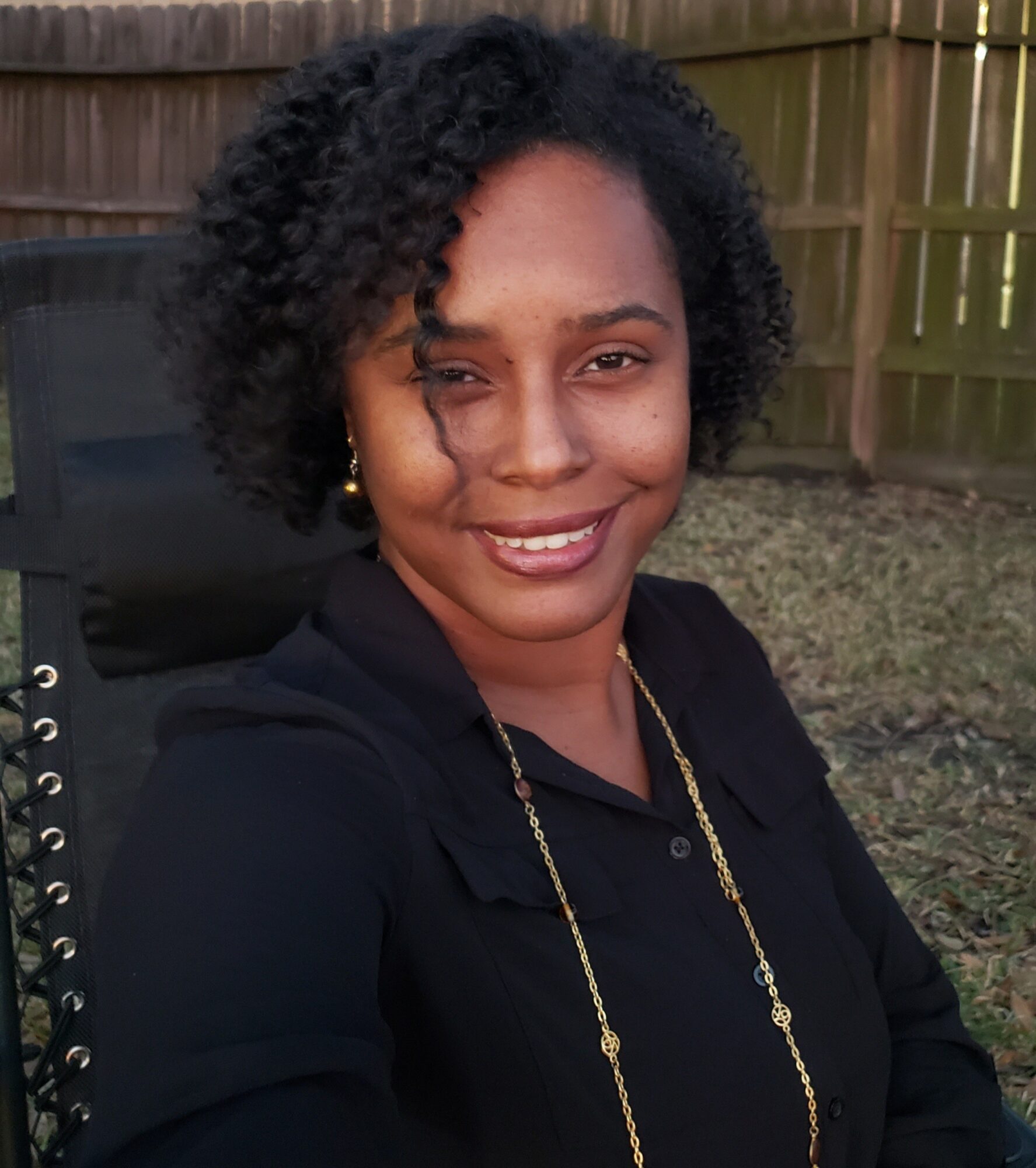 Woman with curly hair smiling outdoors, wearing a black top and gold necklace, in a backyard with a wooden fence.