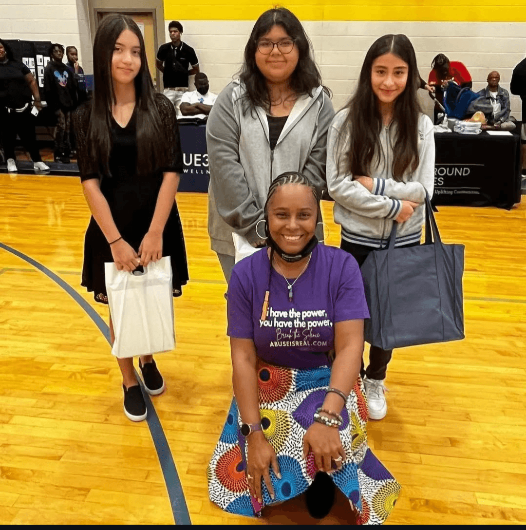 Four girls in a gymnasium, three standing and one kneeling, smiling, with people and equipment in the background.