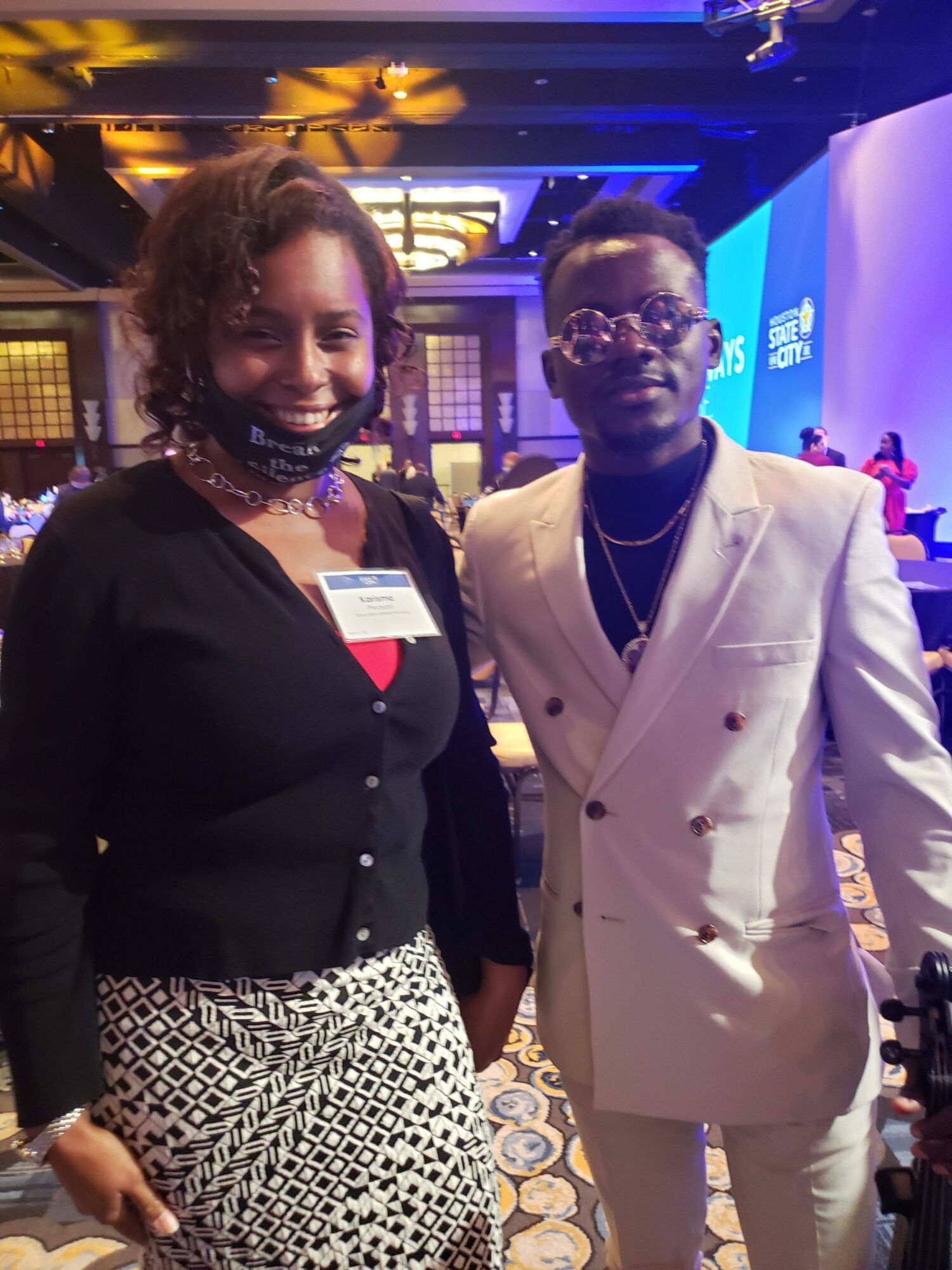 Two people smiling at an indoor event, one wearing a white blazer and the other in a black top with patterned pants.
