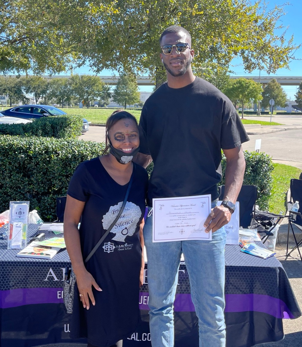 Two people standing outdoors, one holding a certificate, smiling, with trees and a table in the background.