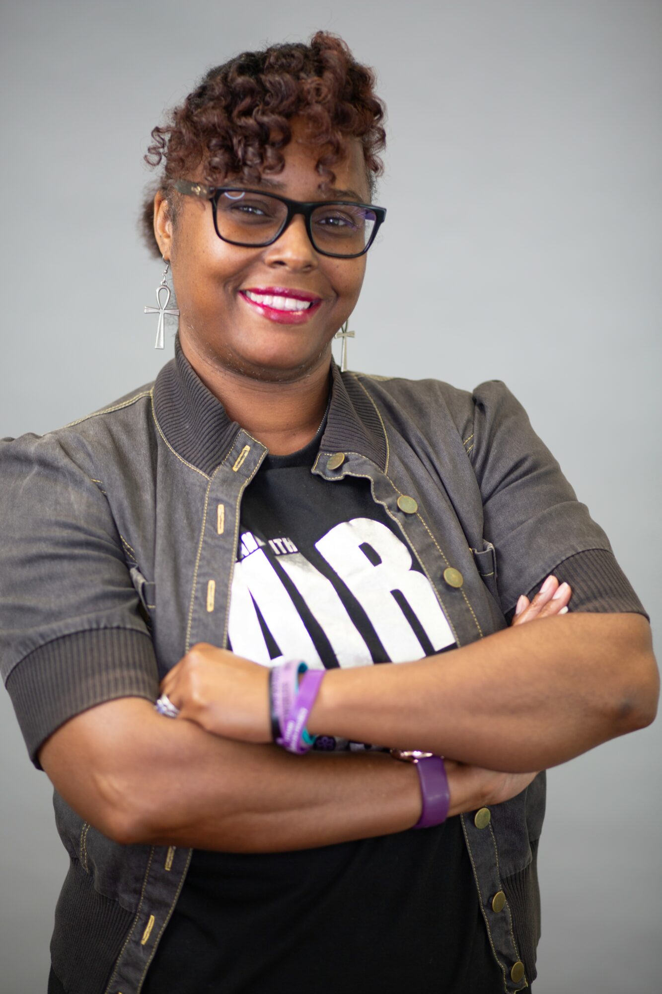Woman with short curly hair, glasses, smiling, arms crossed, wearing a black shirt and jacket, against a plain background.