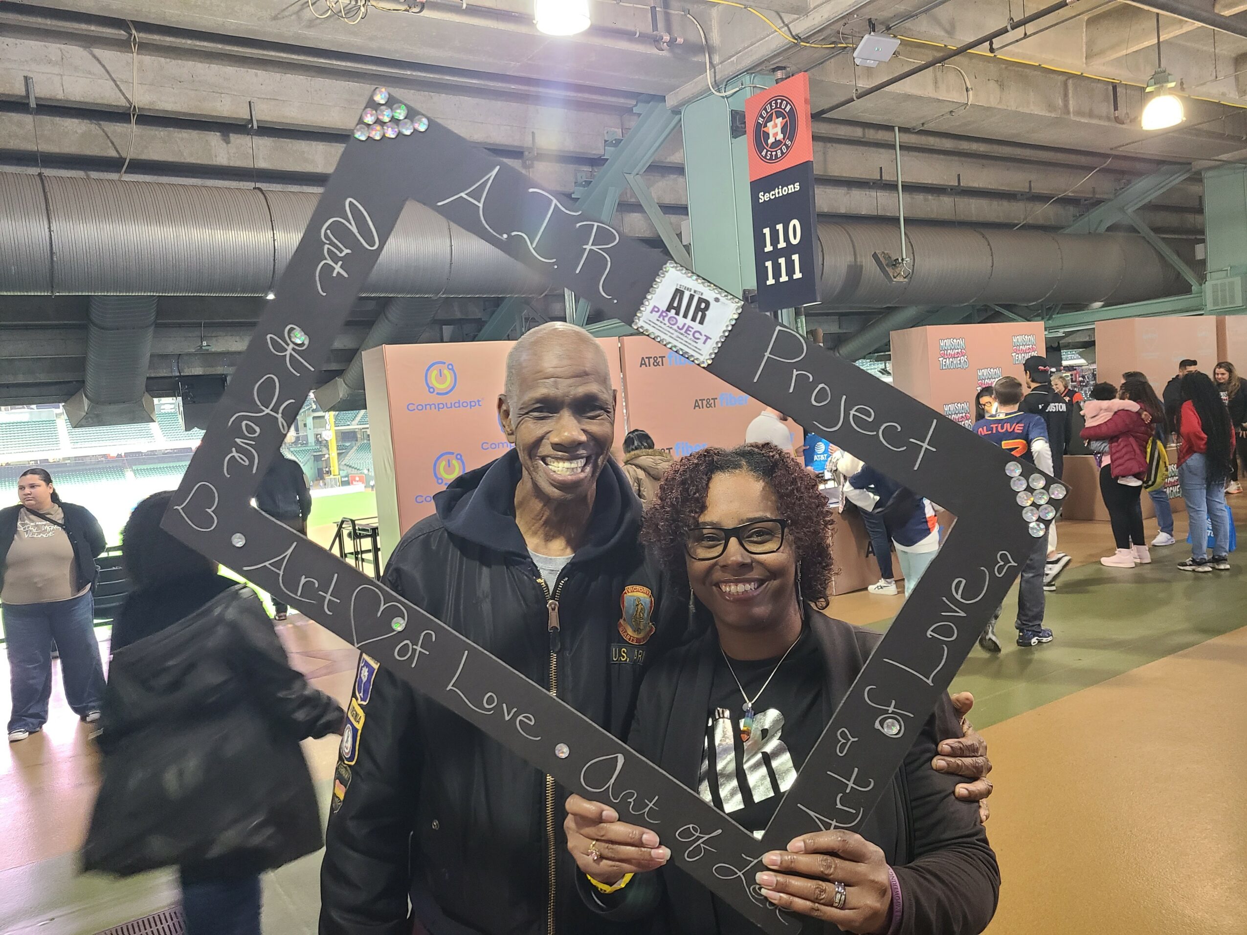 Two smiling people holding a black frame with handwritten text at an indoor event, with other attendees in the background.