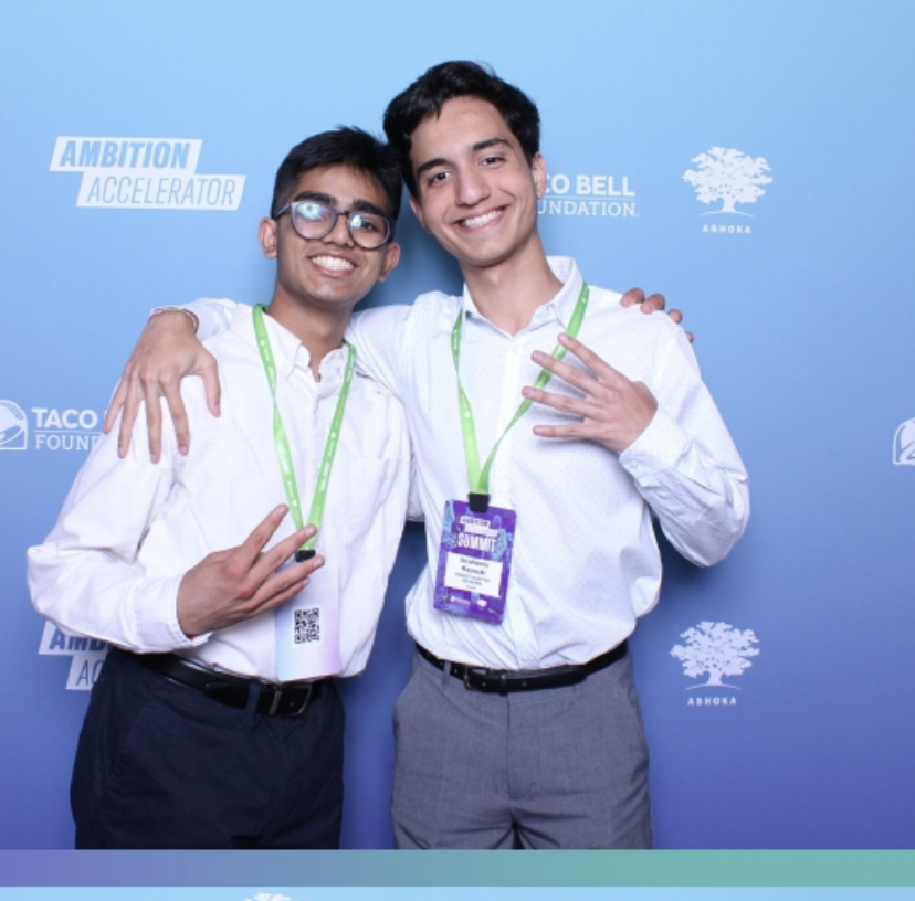 Two young men smiling and posing with arms around each other against a blue background with logos.