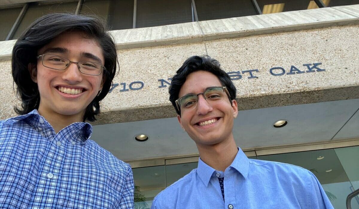 Two young men with glasses smiling outdoors in front of a building with partial text visible.