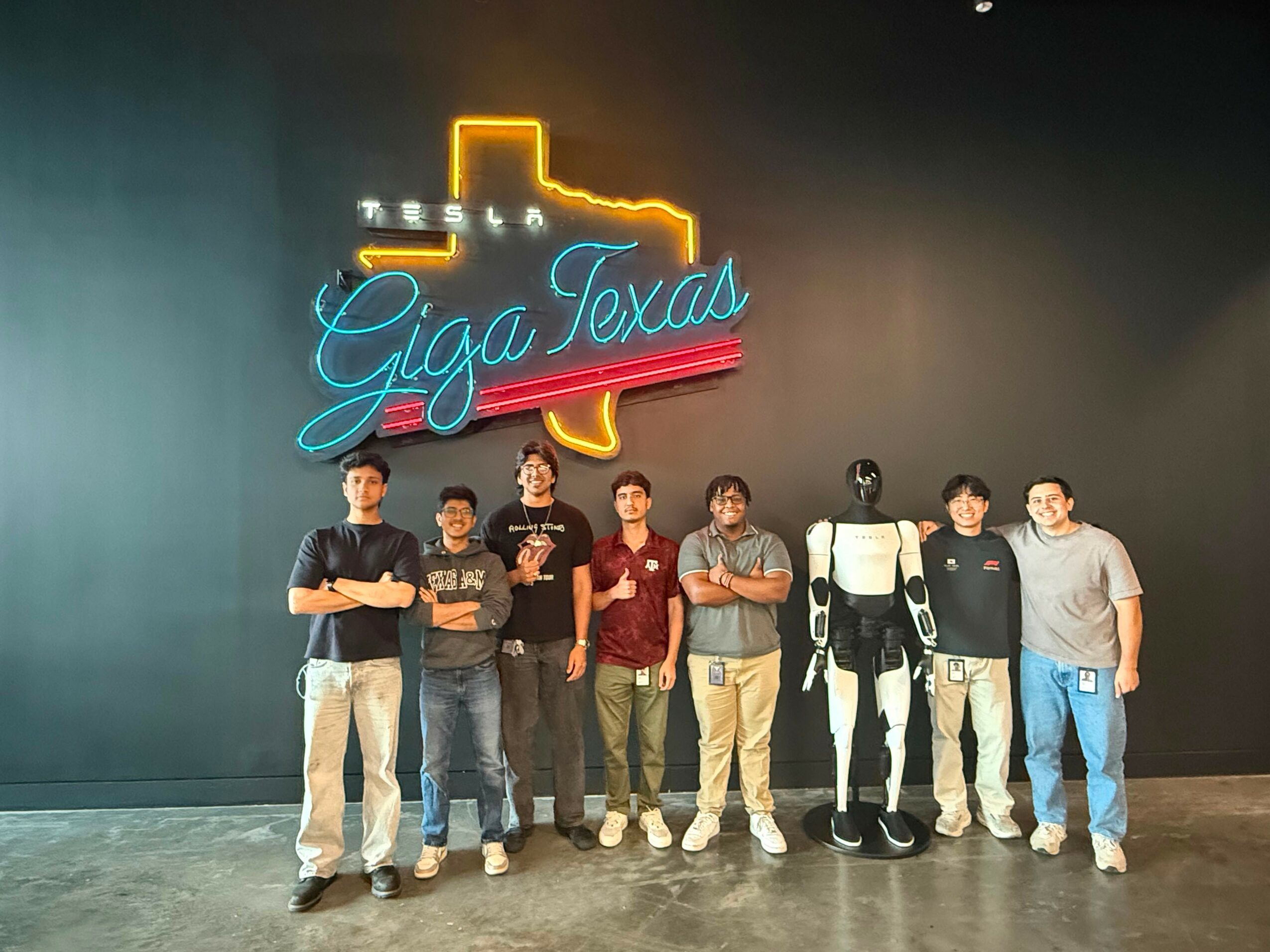 Group of seven people and a robot figure standing in front of a neon Texas shape and 'Giga Texas' sign.