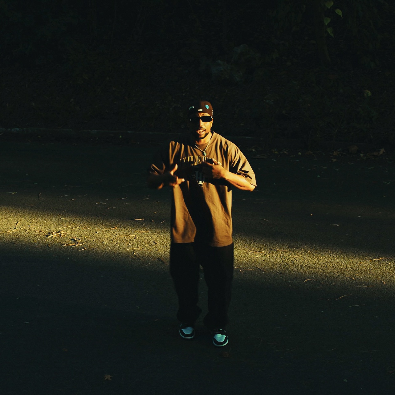 Person standing outdoors in sunlight, wearing sunglasses, a cap, and casual clothing, holding an object, with dark background.