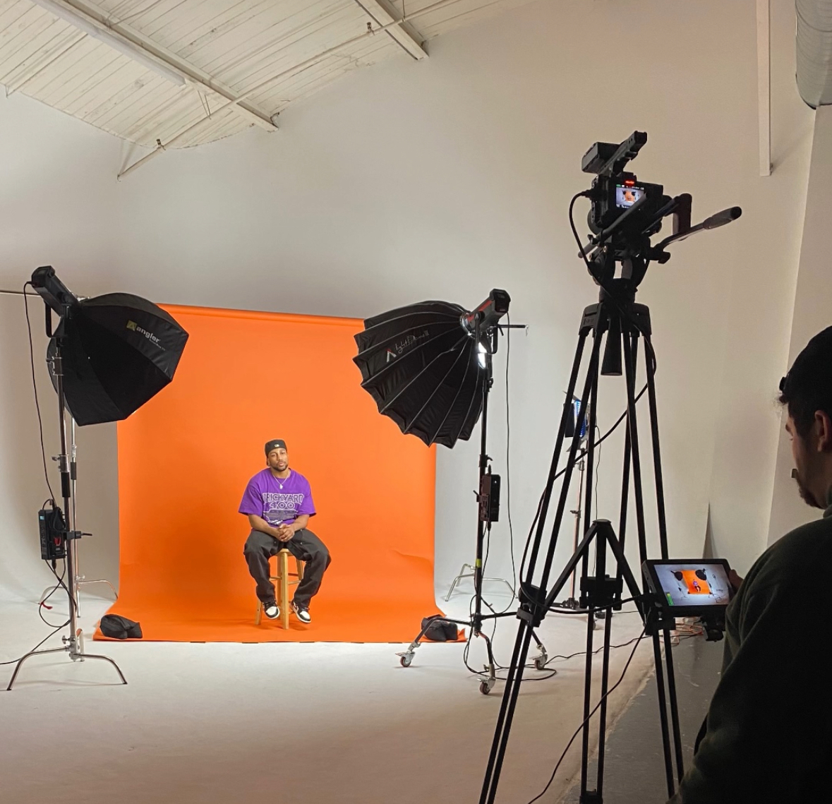 Person sitting on a stool in front of an orange backdrop, surrounded by studio lighting and camera equipment.