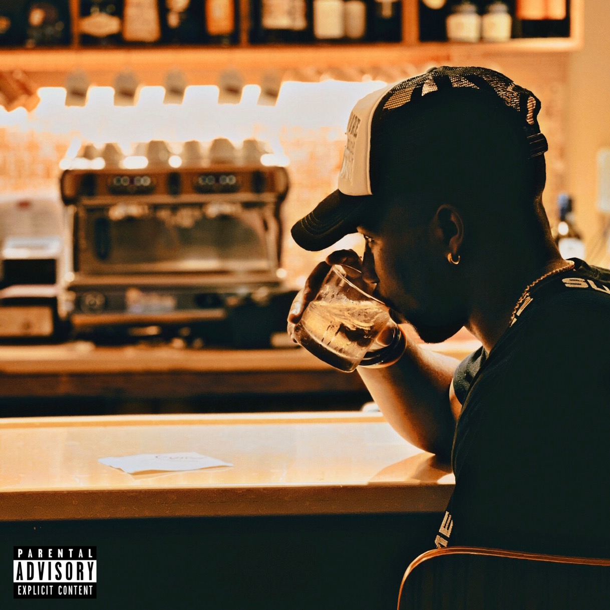 Person wearing a cap drinks from a glass at a bar counter, with a blurred background of bottles and equipment.