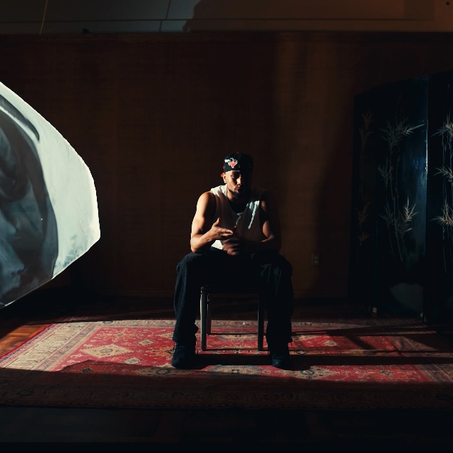 Person sitting on a chair in a dimly lit room, holding a book, with a patterned rug and a dark background.