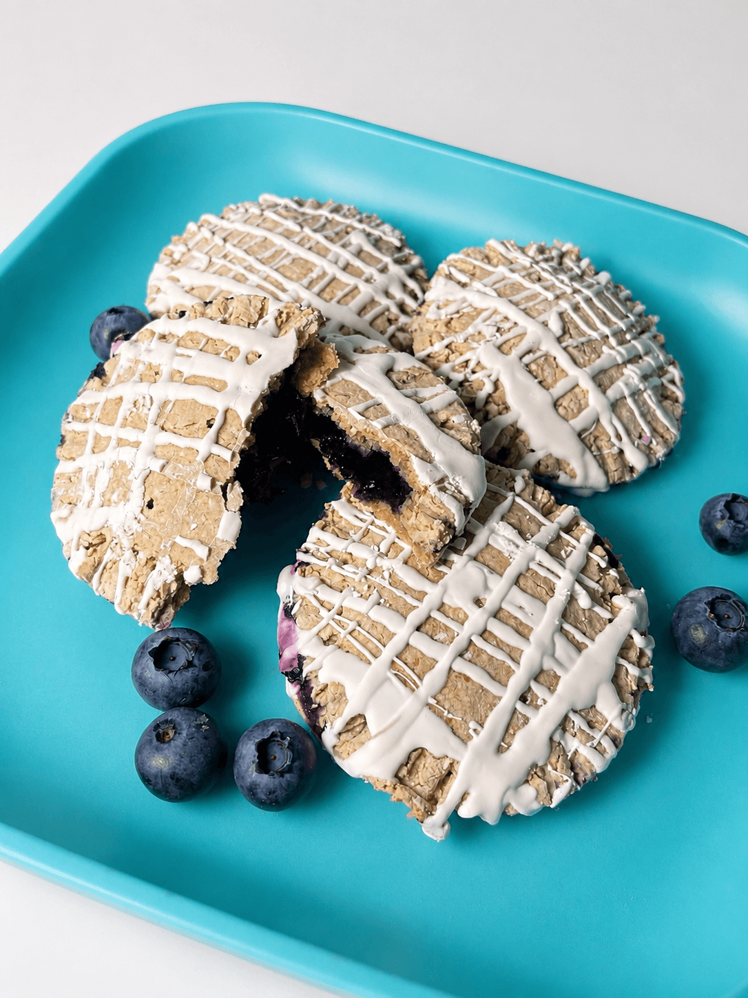 Four cookies with white icing and blueberries on a turquoise plate, one cookie broken showing dark filling inside.