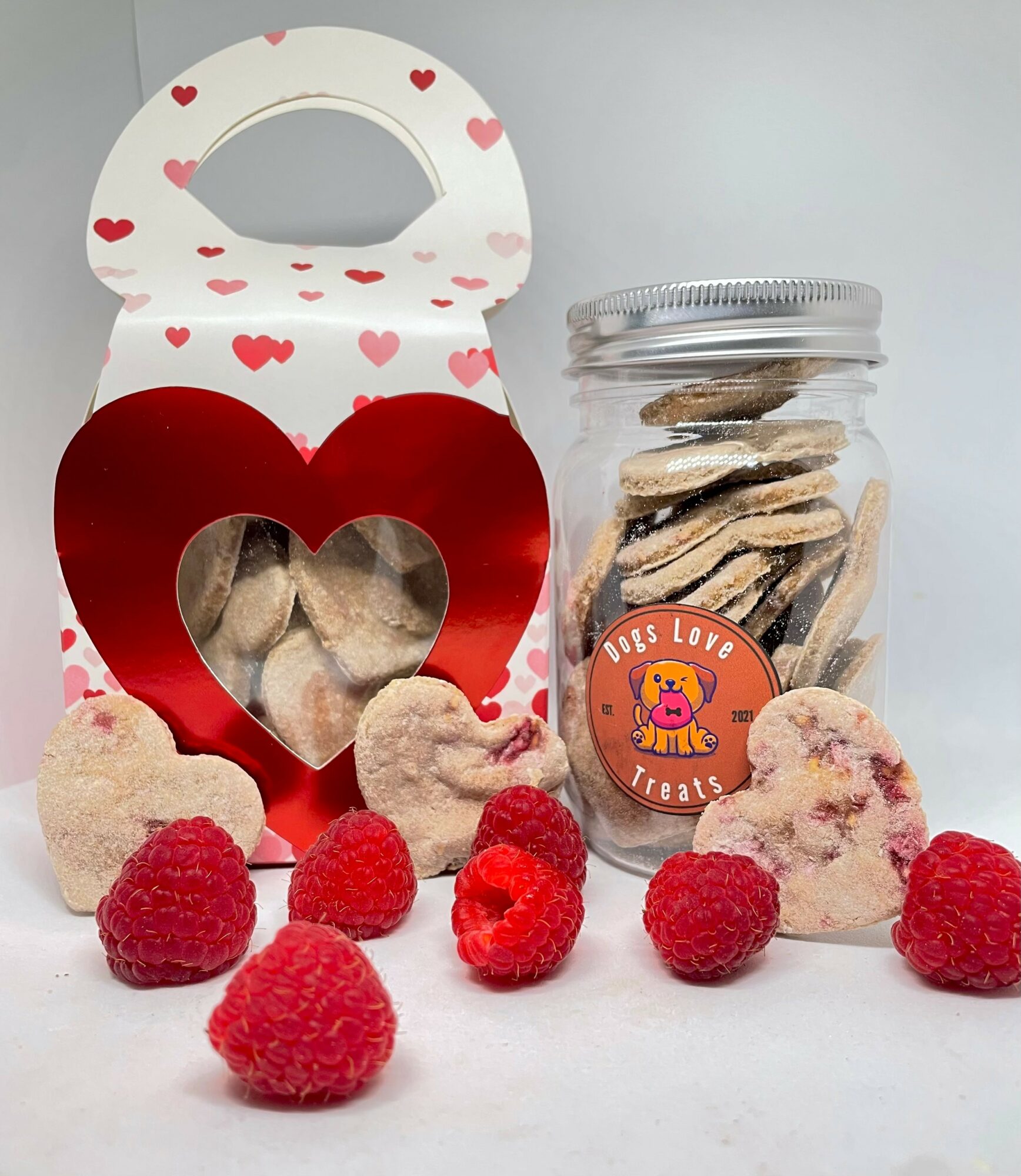 Heart-shaped box with a clear window, jar of cookies, and raspberries on a white surface.