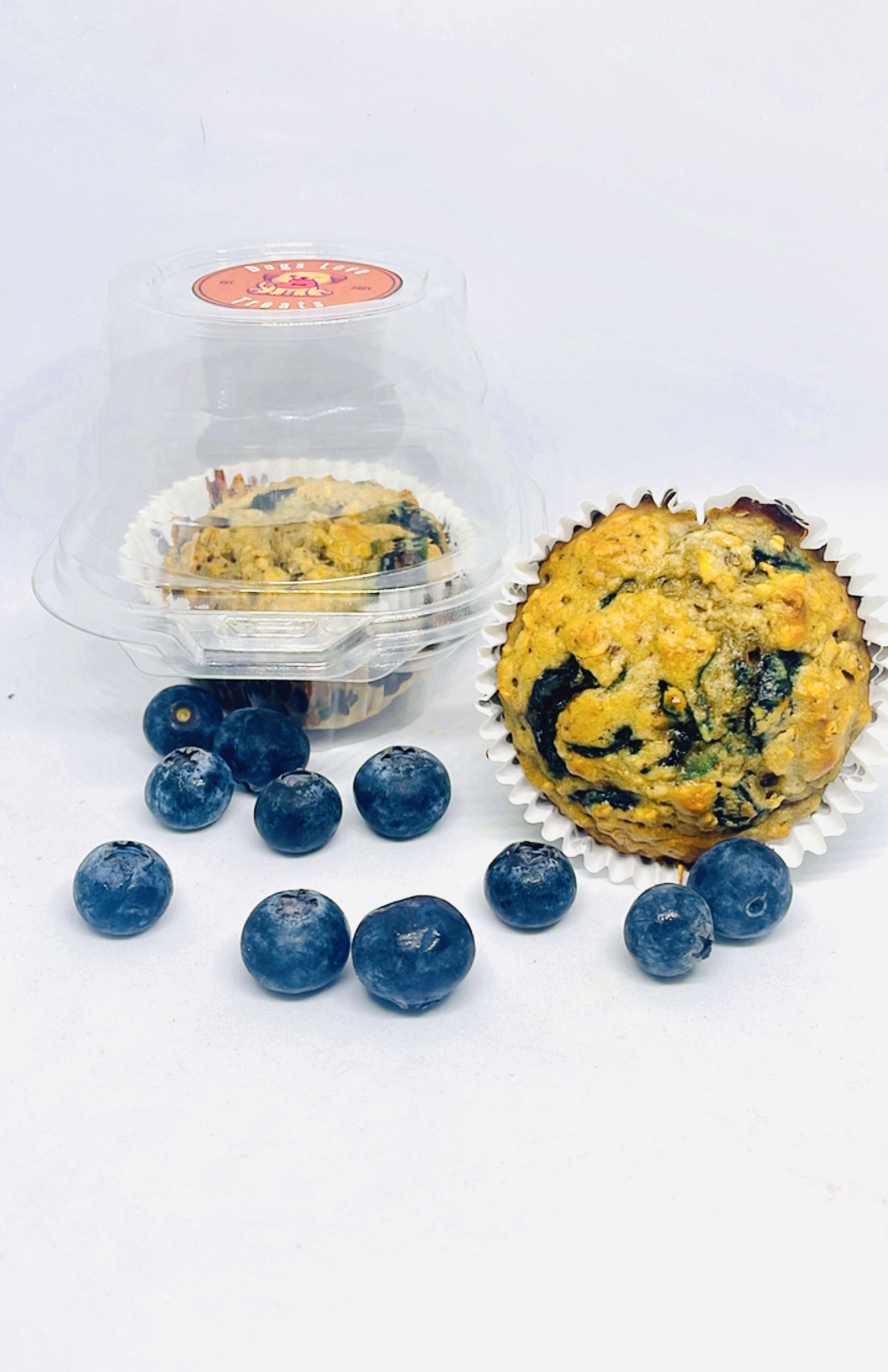 Blueberries scattered around a yellow muffin with dark spots, with a clear plastic container in the background.