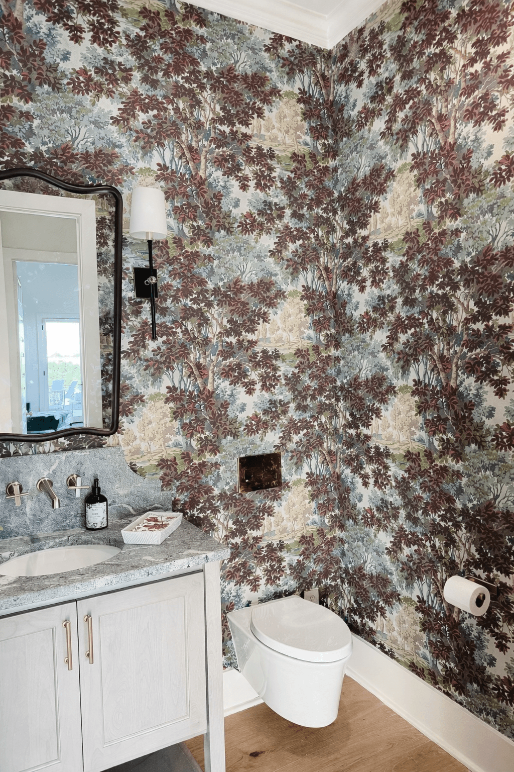 Bathroom corner with floral wallpaper, mirror, wall lamp, sink, toilet, and toilet paper holder.