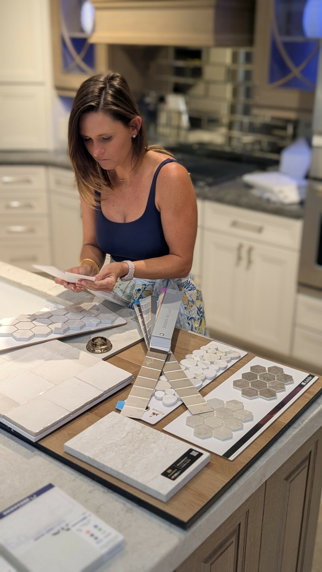Woman in blue top examining tile samples on kitchen counter, with various tile options displayed.