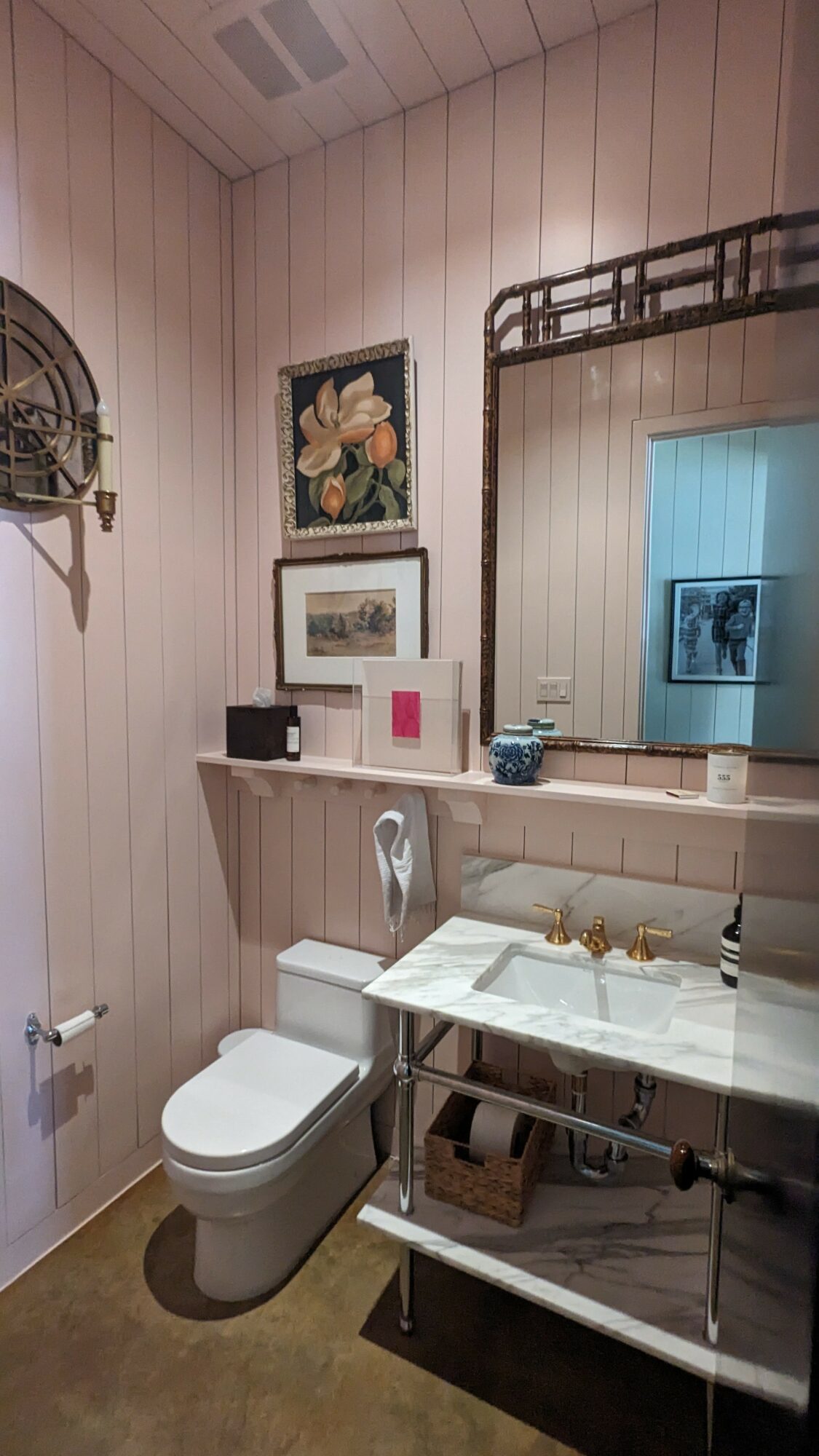 Bathroom with toilet, mirror, wall art, and a shelf with items, beige walls, and a window with a view outside.