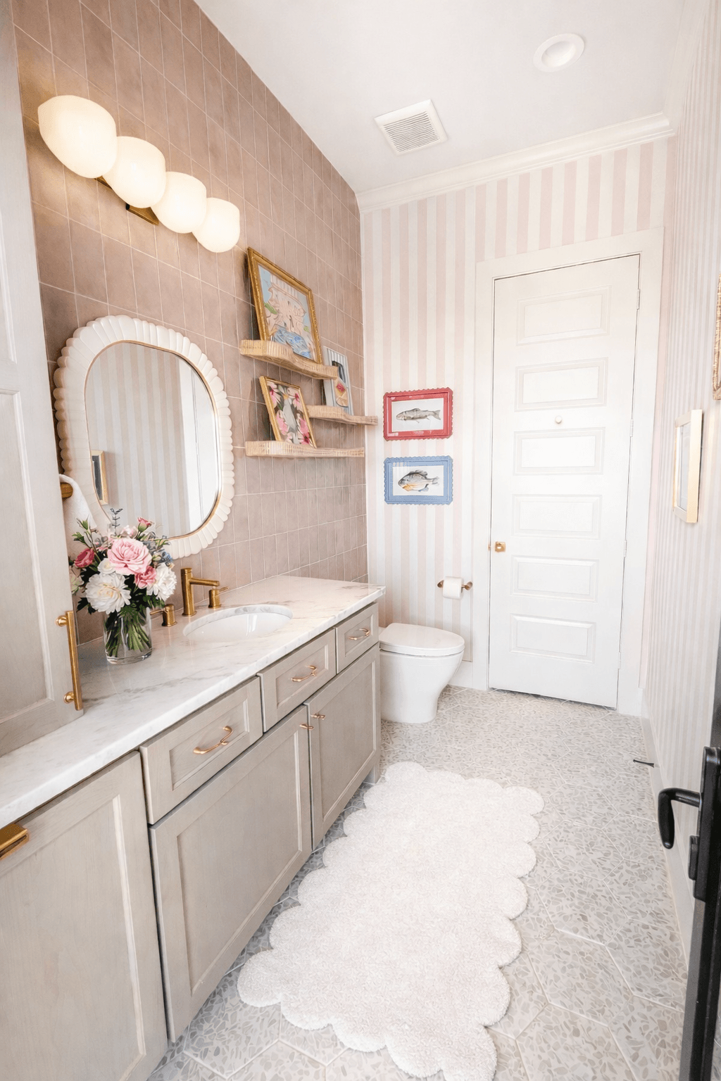 Bathroom with beige vanity, mirror, flowers, framed pictures, toilet, white door, pink striped wall, and fluffy white rug.