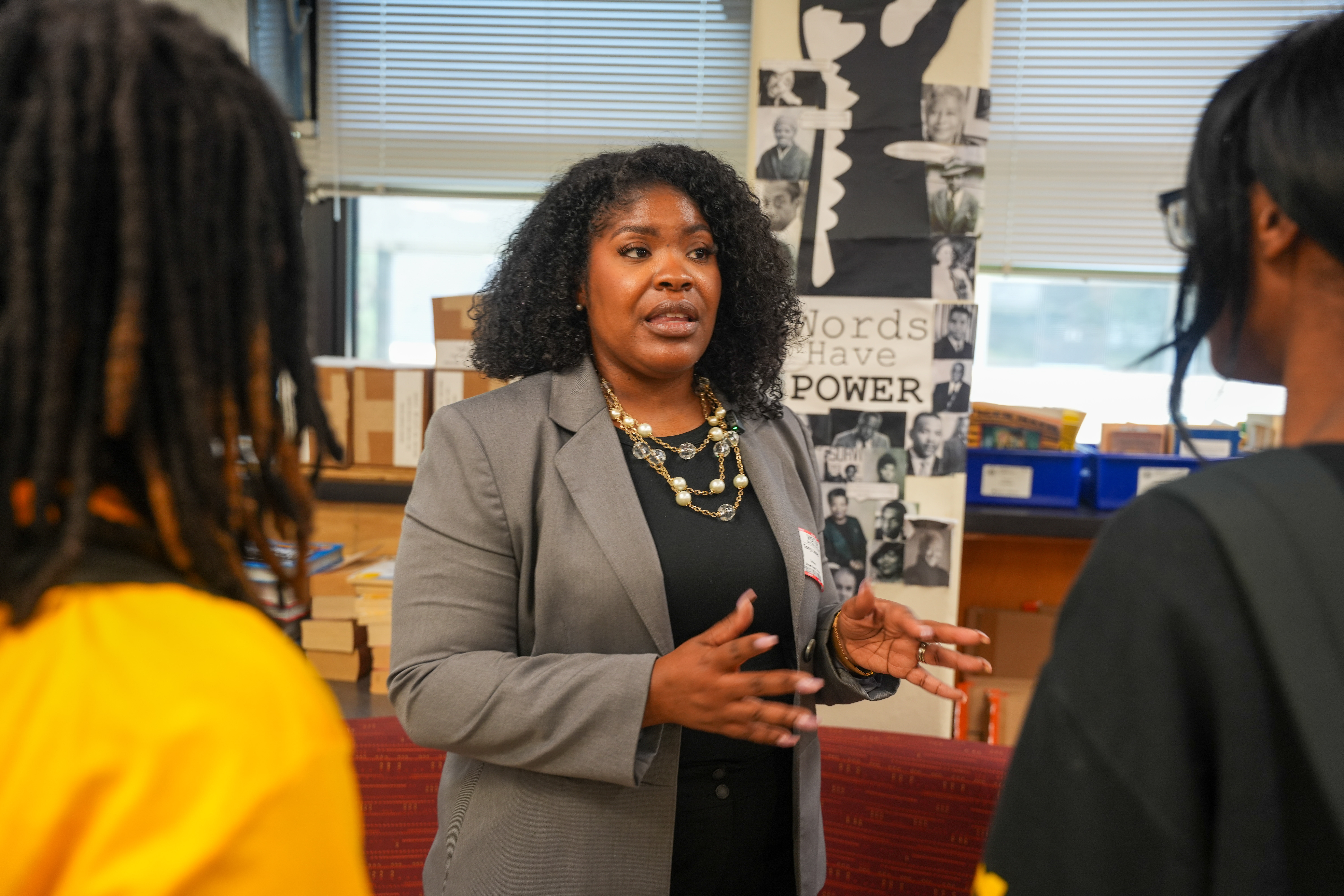 Woman speaking to two people in a classroom or office setting, with posters and books in the background.