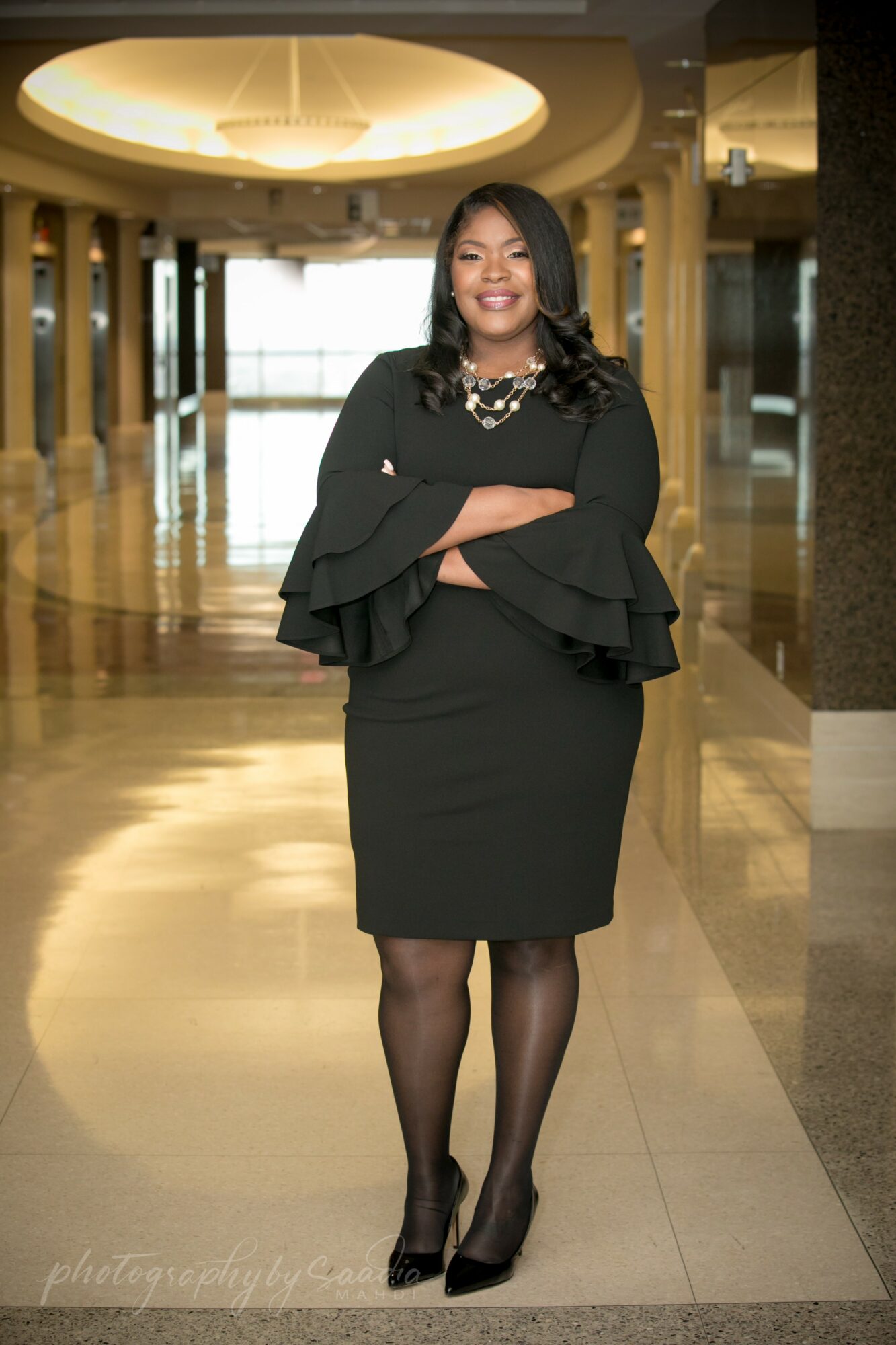 Woman in black dress with ruffled sleeves standing in a spacious, well-lit hallway with a circular ceiling light.