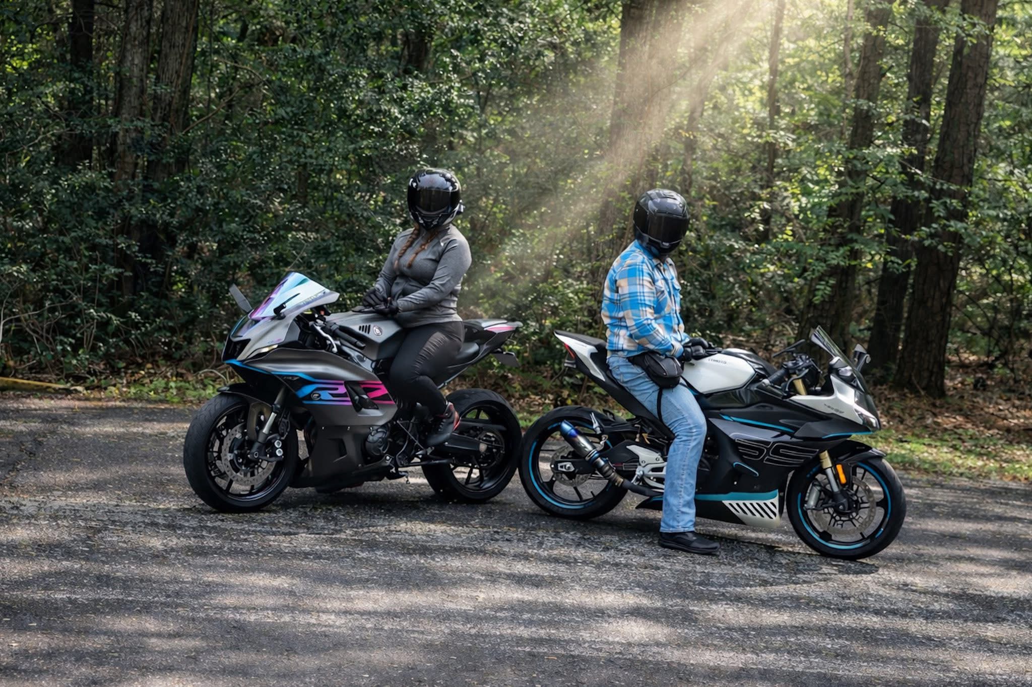Two people on motorcycles parked on a forest road with sunlight filtering through trees.
